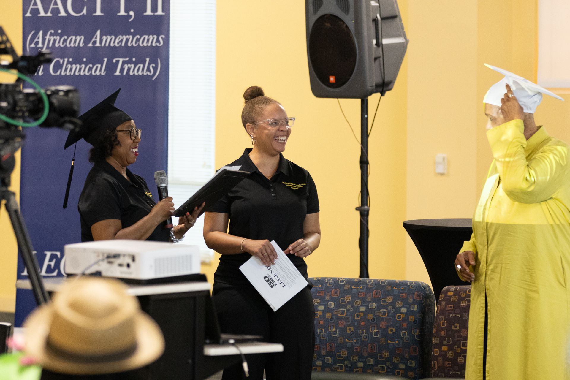 Two women in black, one in graduation cap, present diploma to person in yellow robe.