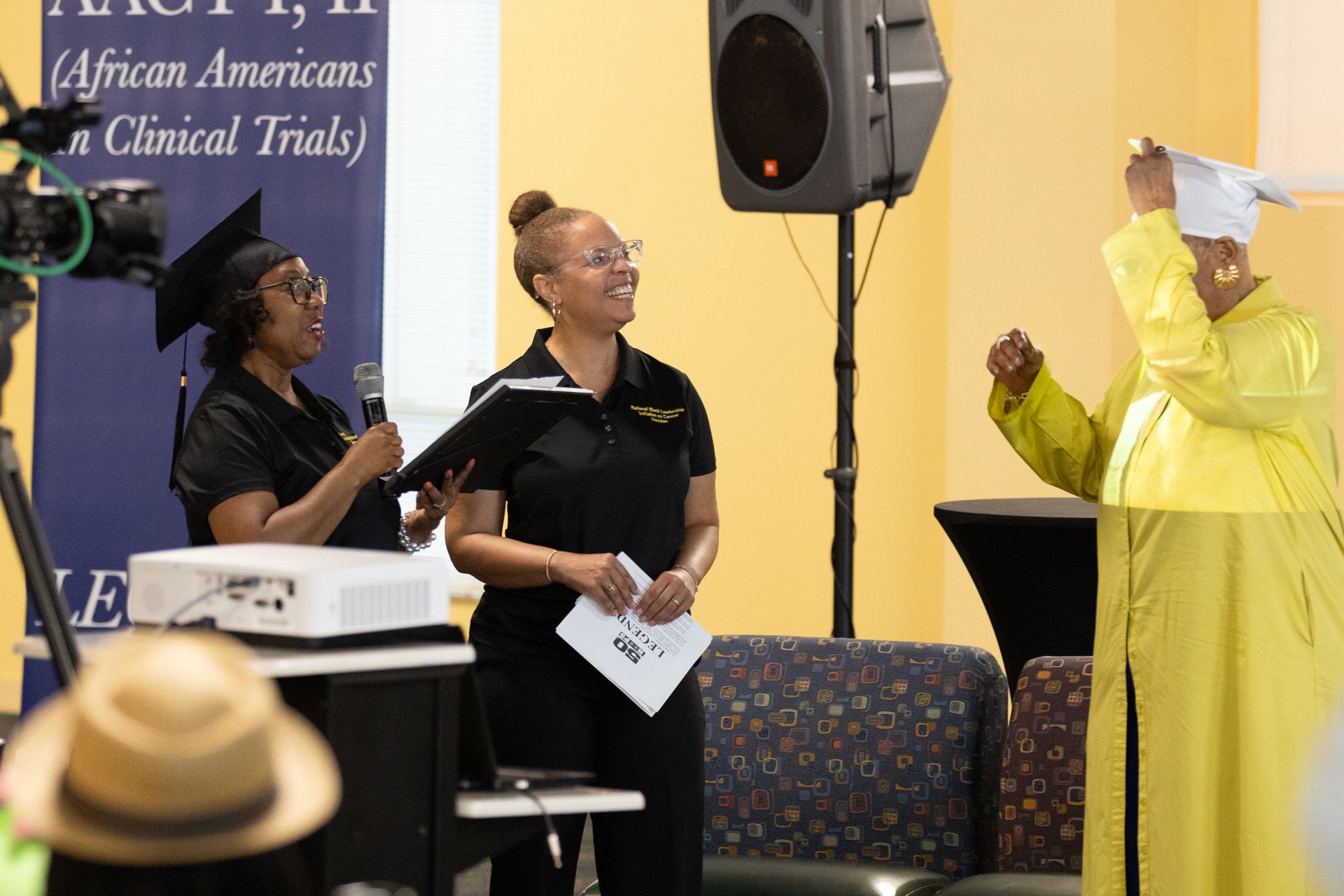 Two women at a podium, one in cap and gown, smiling at the other. Event is indoors.