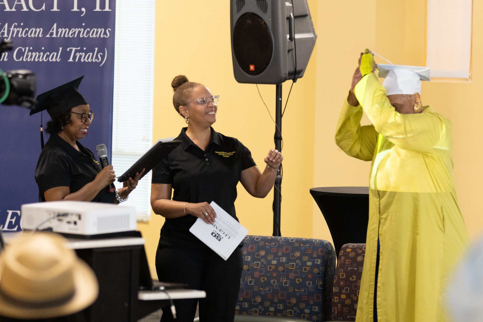 Three people at a podium, one in yellow graduation attire.  A woman adjusts another's mortarboard.  Indoor setting.