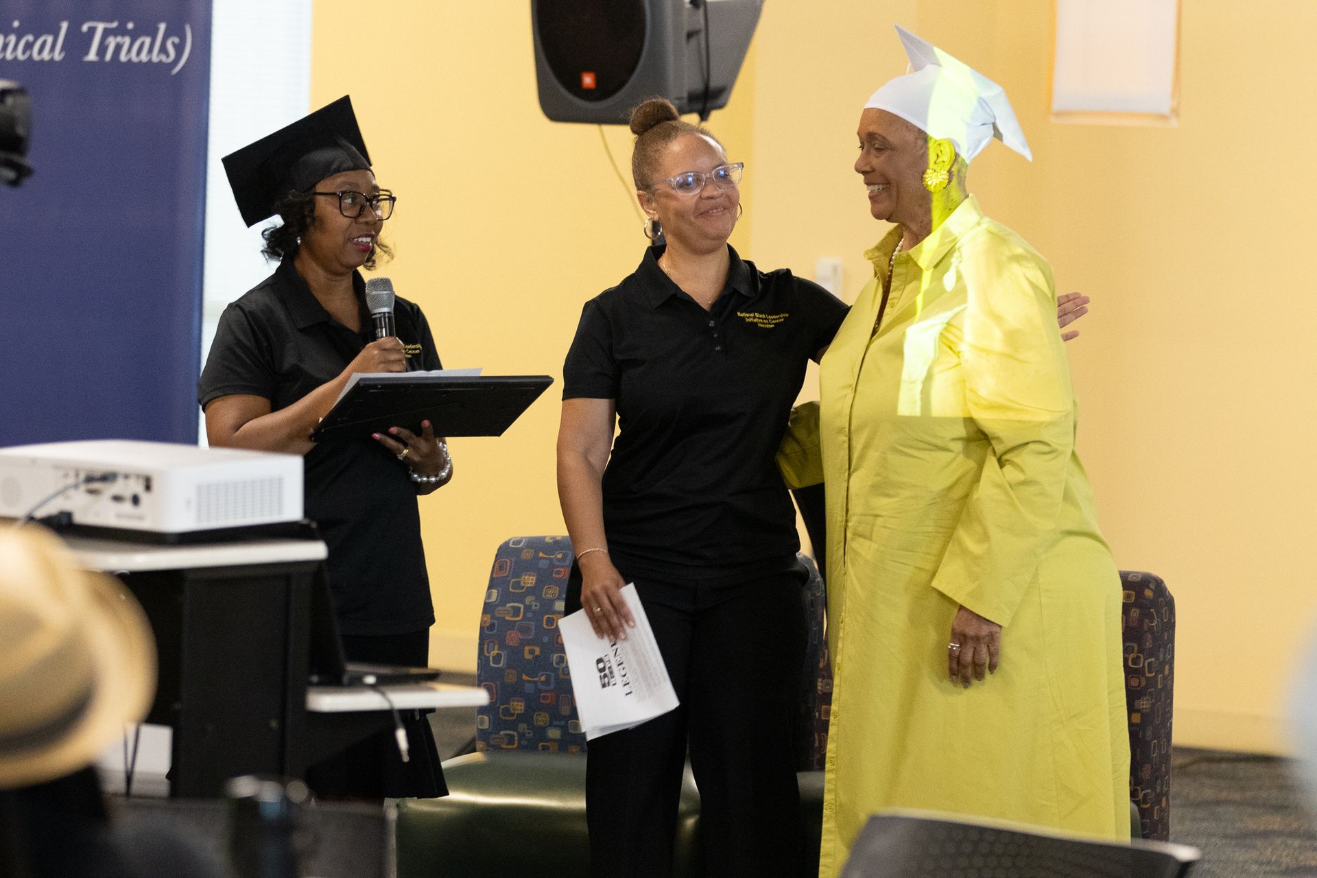Woman in cap and gown with two women in black shirts on a stage.