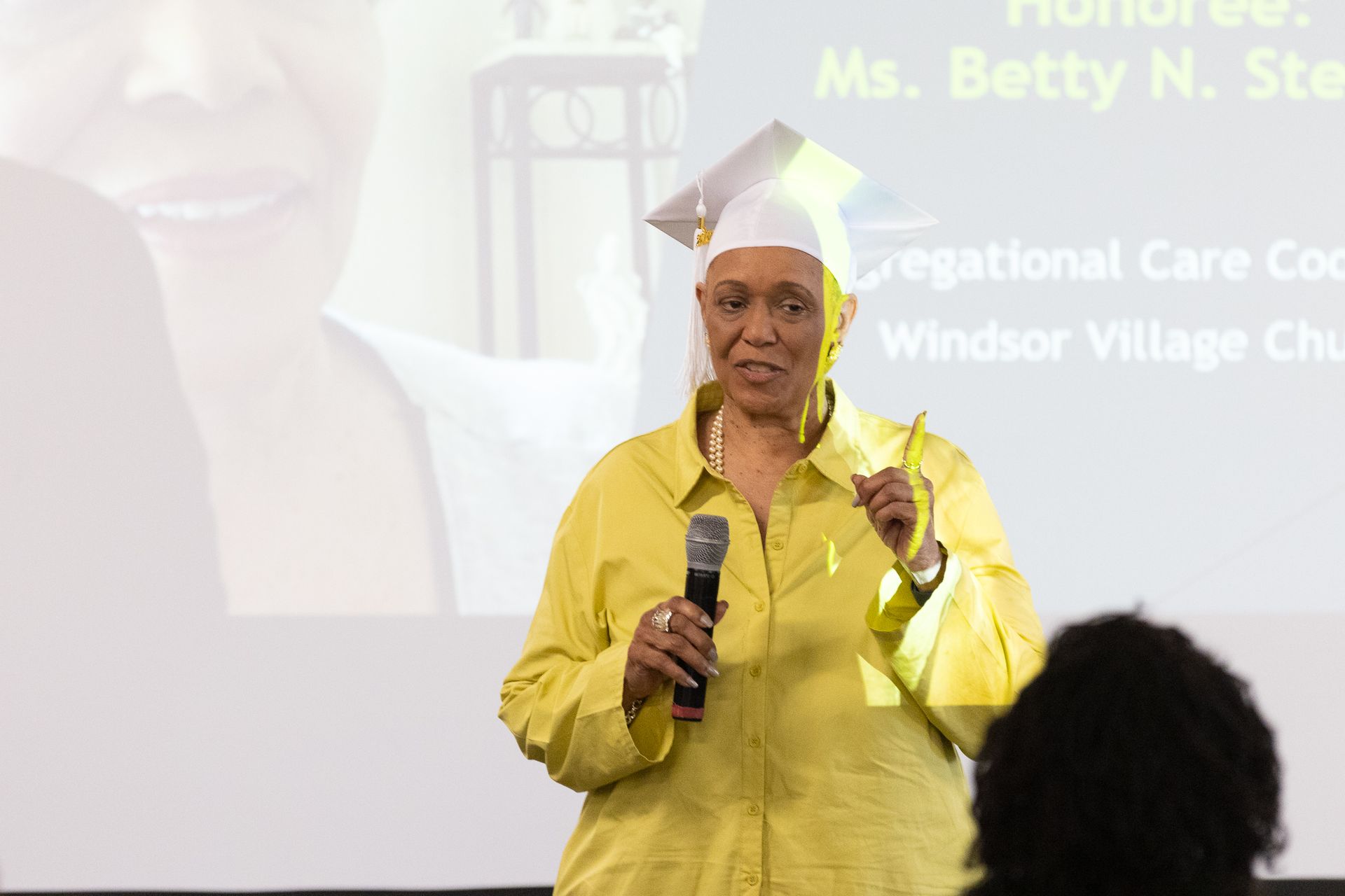 Woman in yellow top and mortarboard speaks into a microphone at a podium.