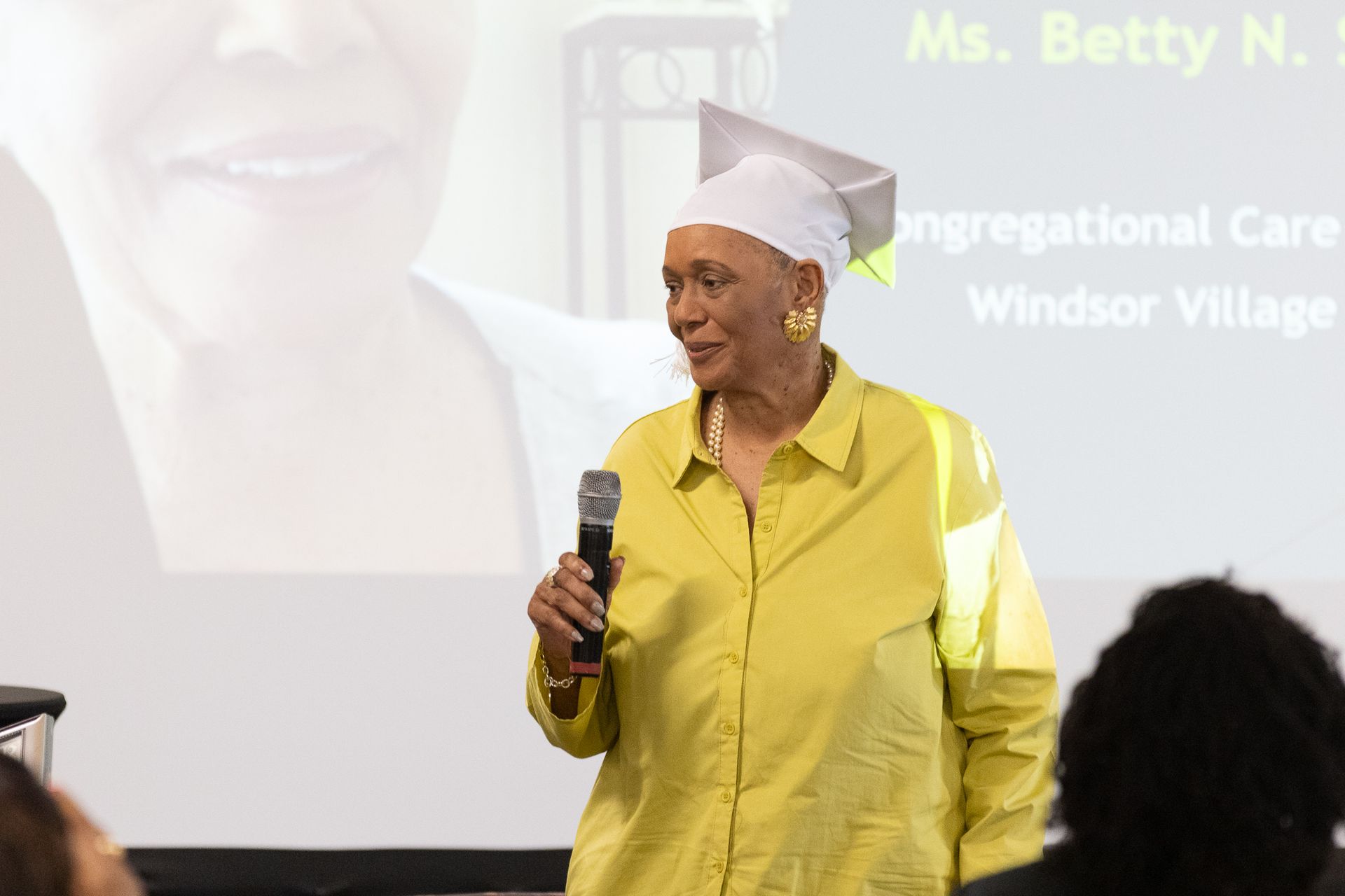Woman in yellow shirt, mortarboard, speaking into a microphone; Windsor Village event.