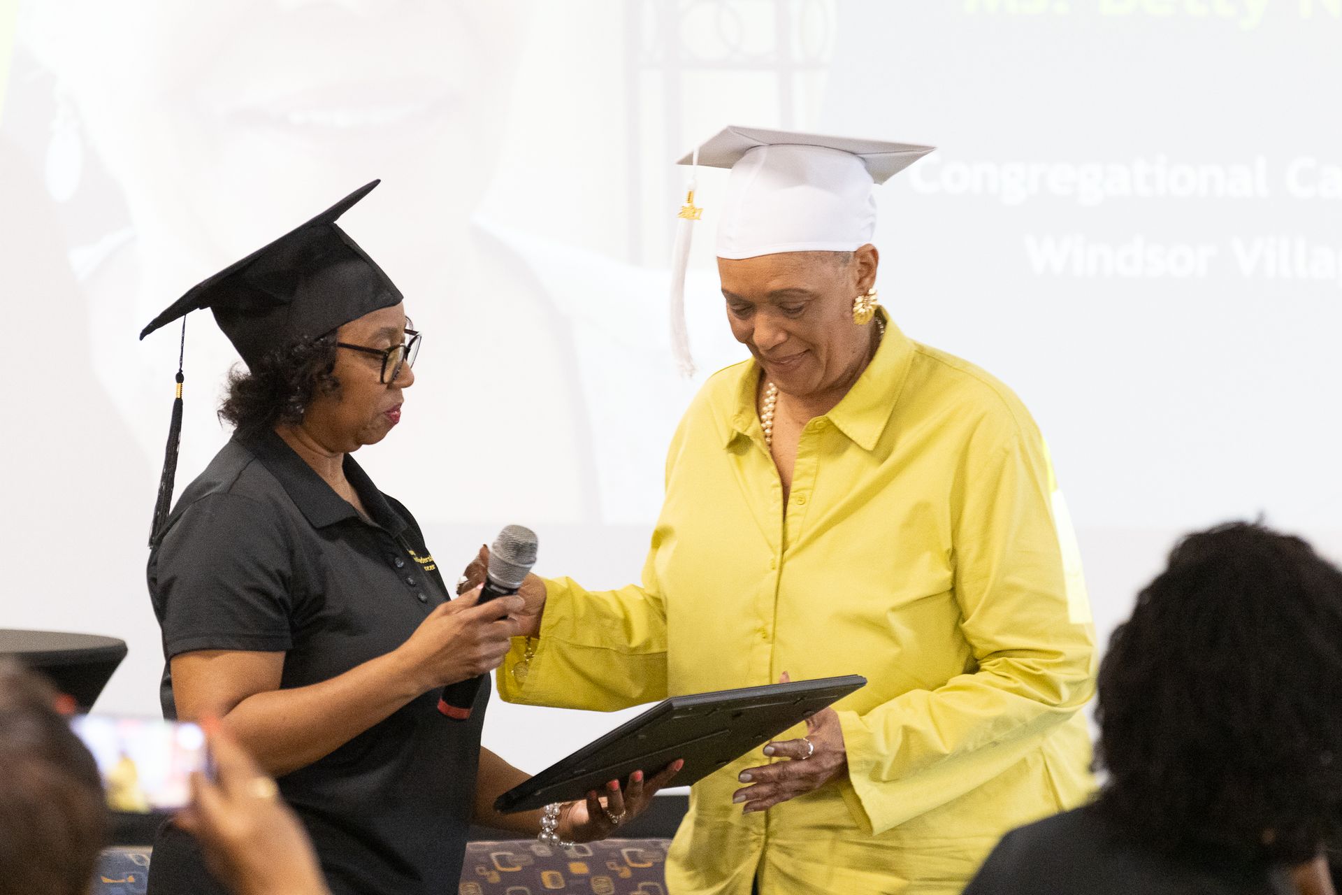 Two women in graduation caps; one presents a diploma at an event.