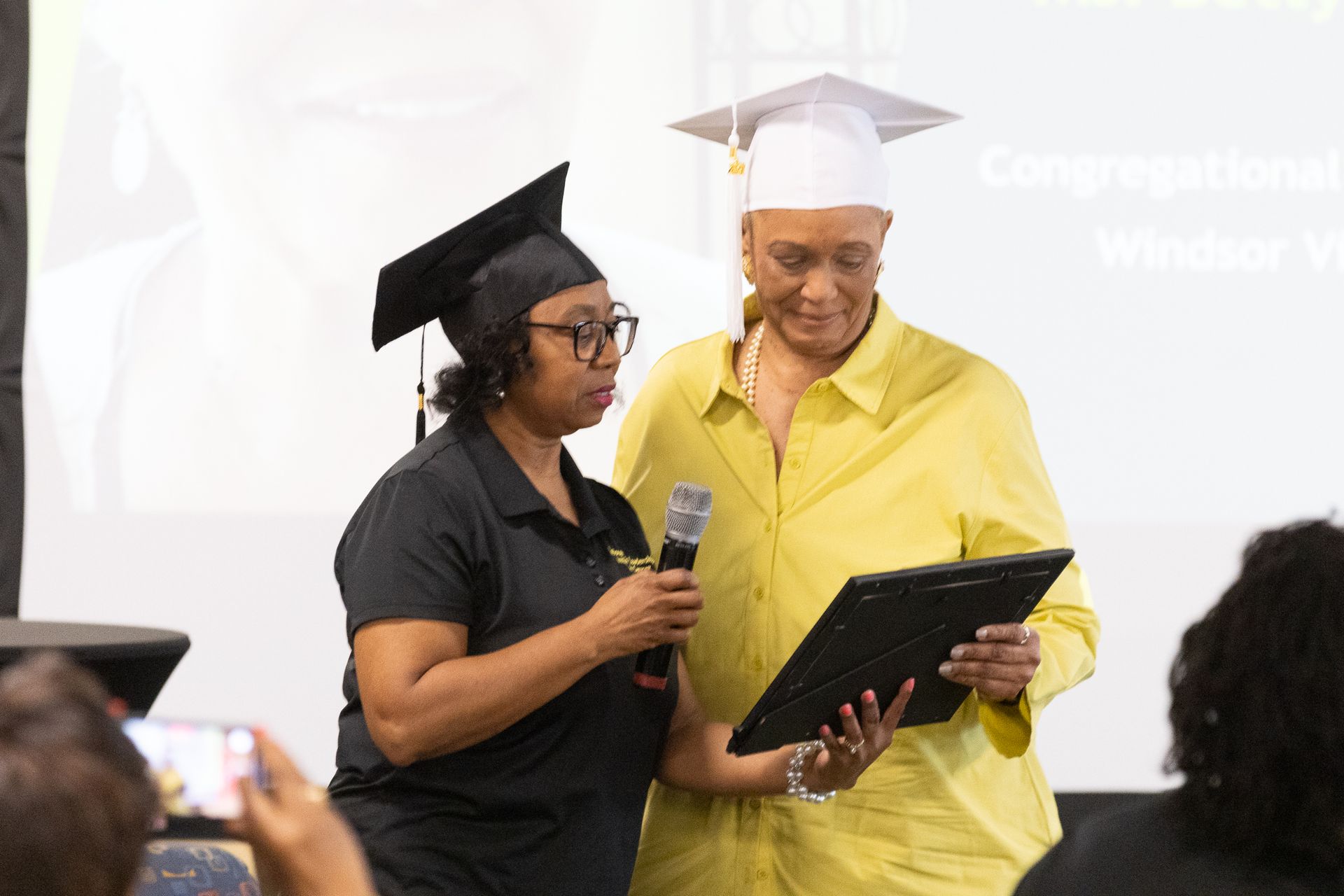 Woman presents framed certificate to another woman wearing graduation cap, both standing at event.