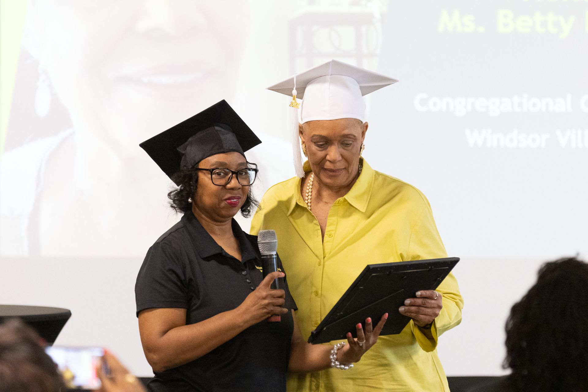 Woman in cap receives award from another at a ceremony, smiling.