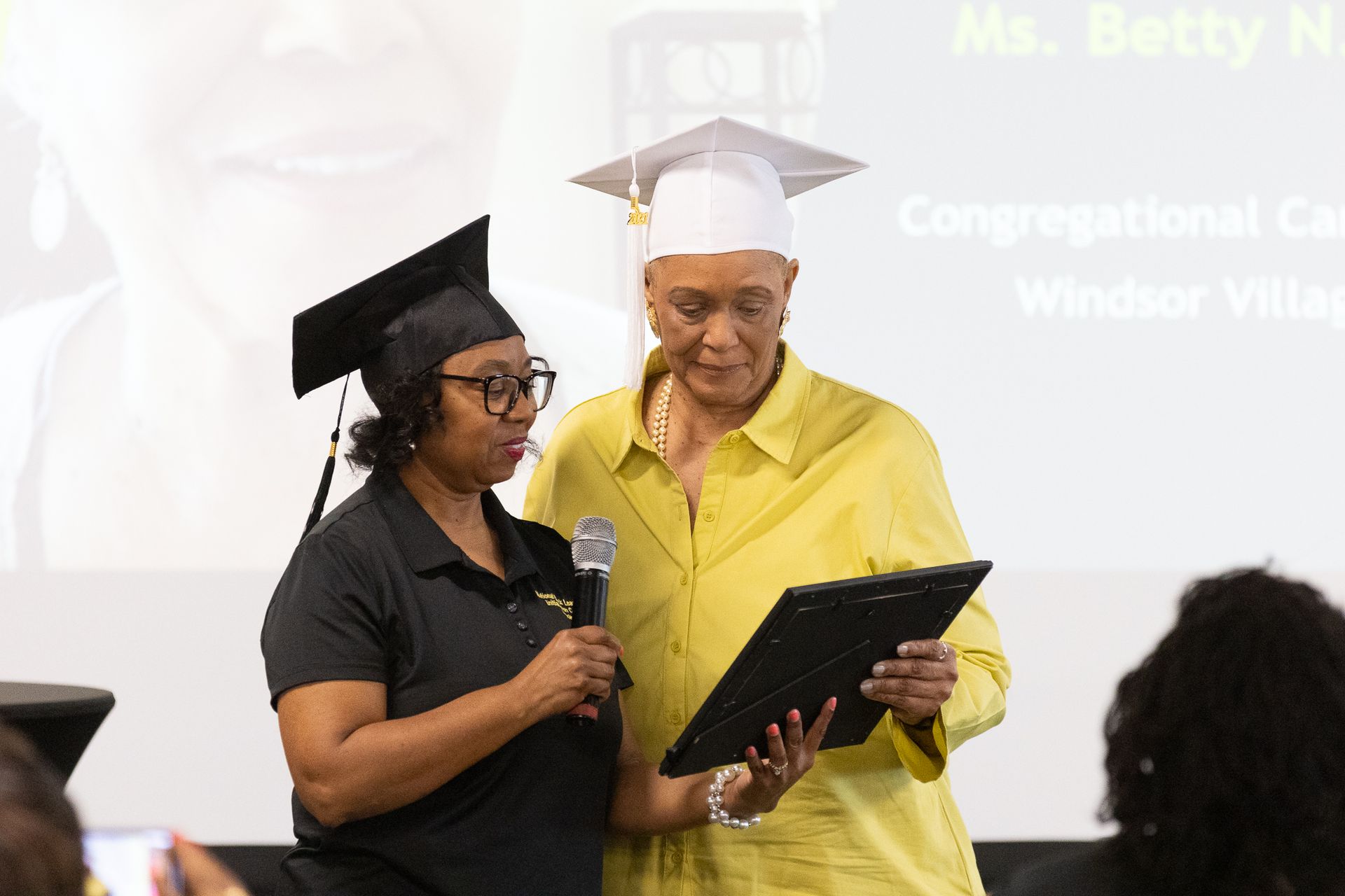 Woman presents award to another, both wearing graduation caps, at a podium.