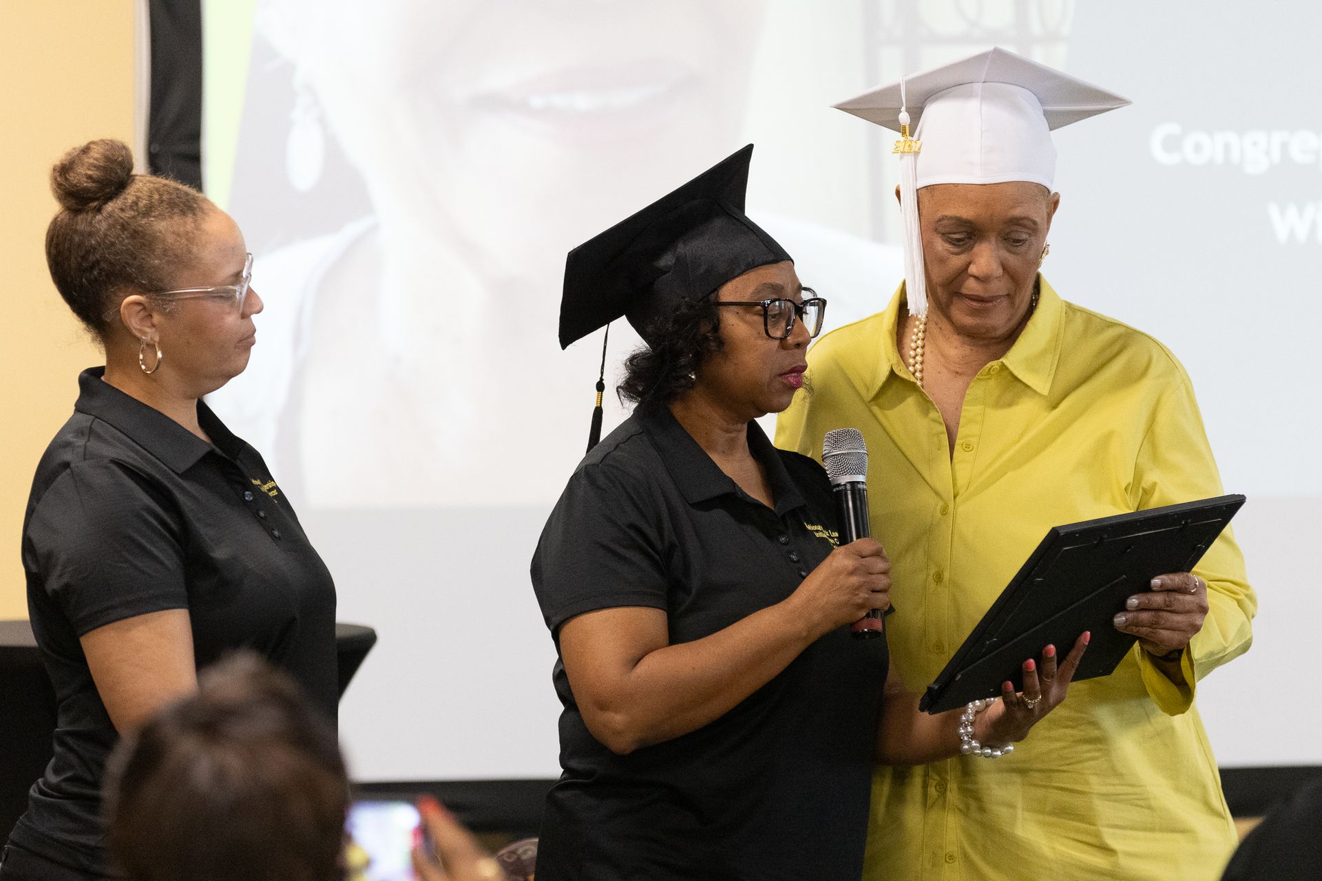 Three women at graduation ceremony: One in a cap speaks into a mic, another presents a certificate.