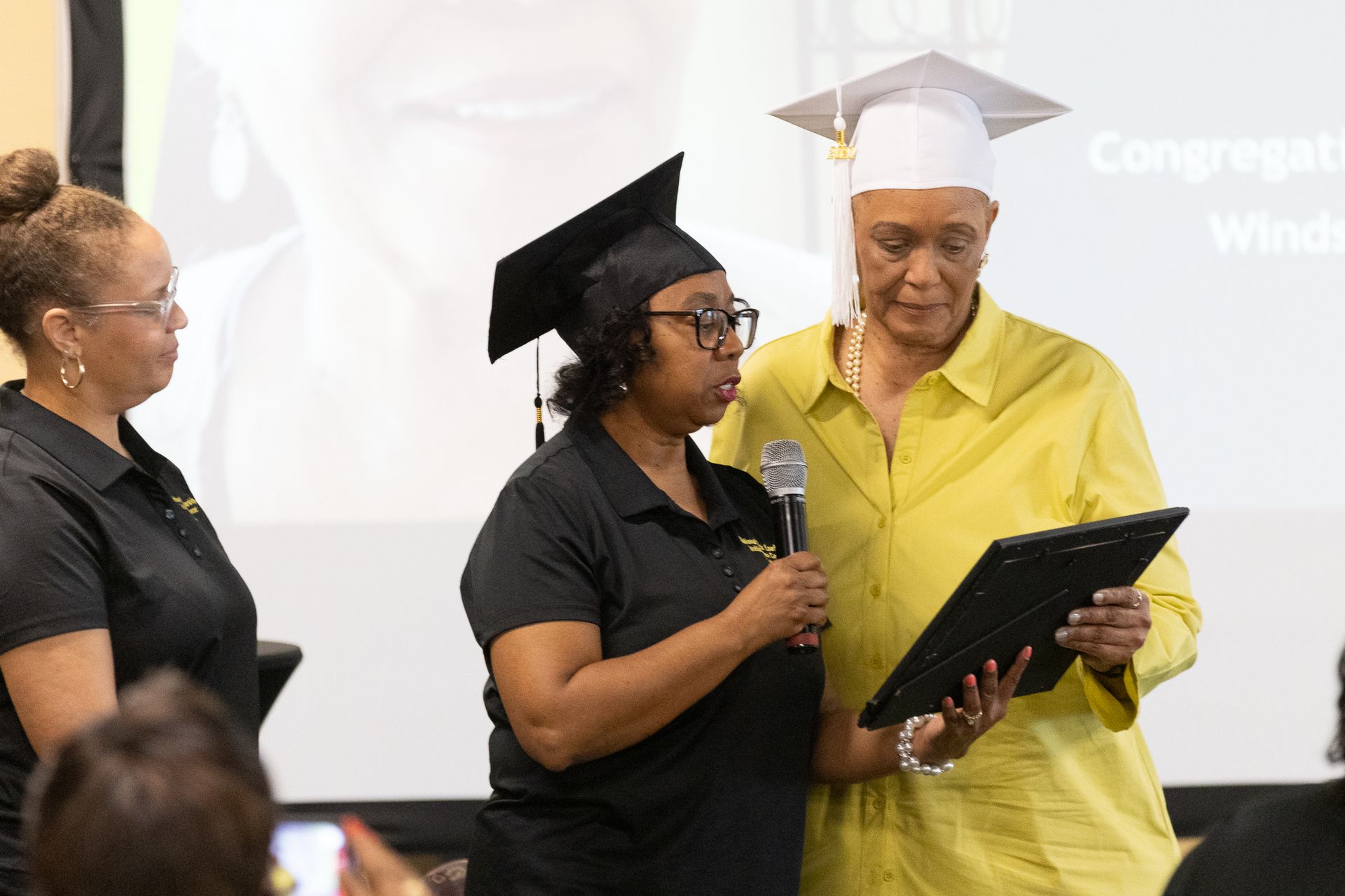 Woman in graduation cap and gown being presented an award by another woman. A third woman watches.