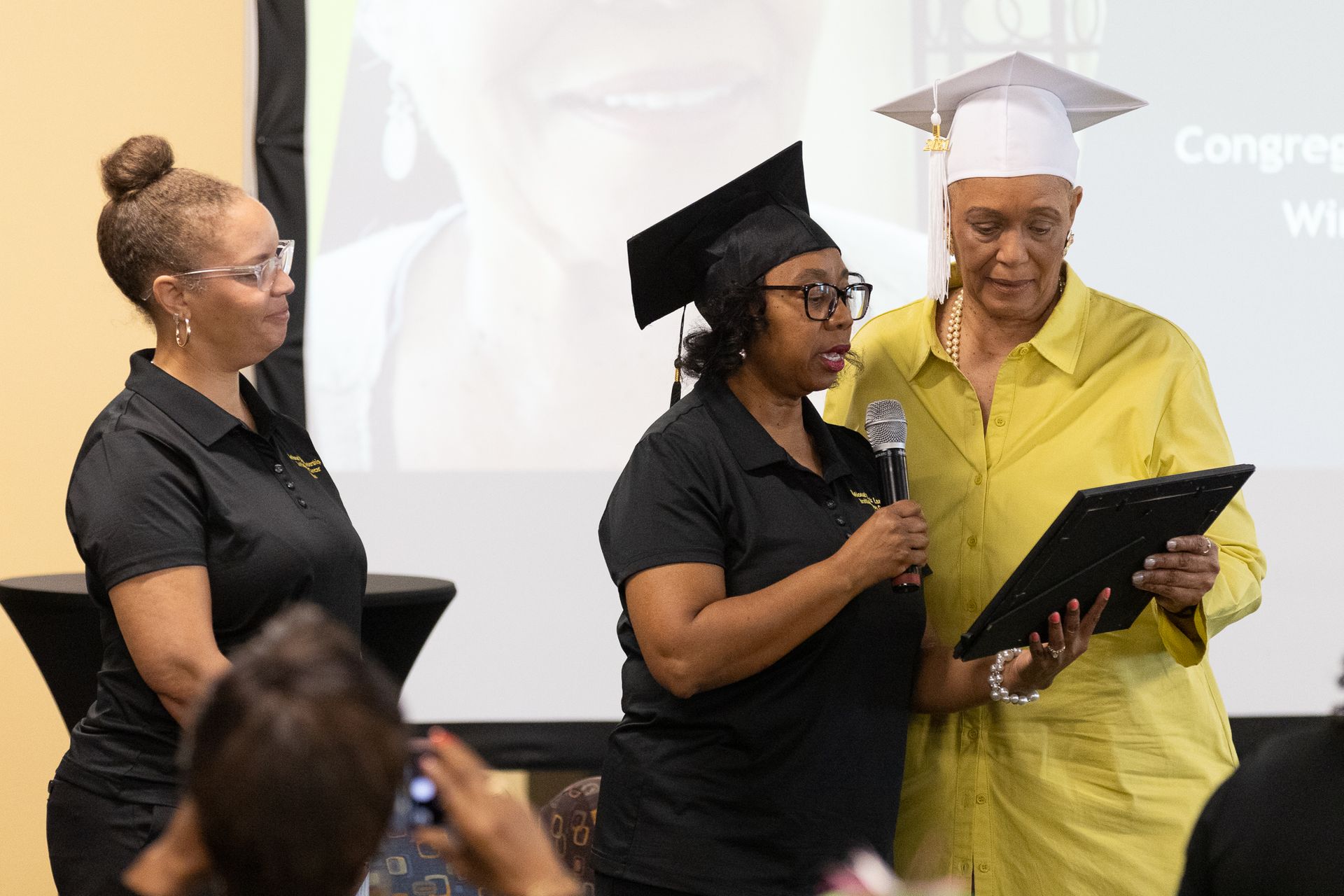 Three people at graduation. One in yellow holding a diploma, a woman speaks into a microphone.