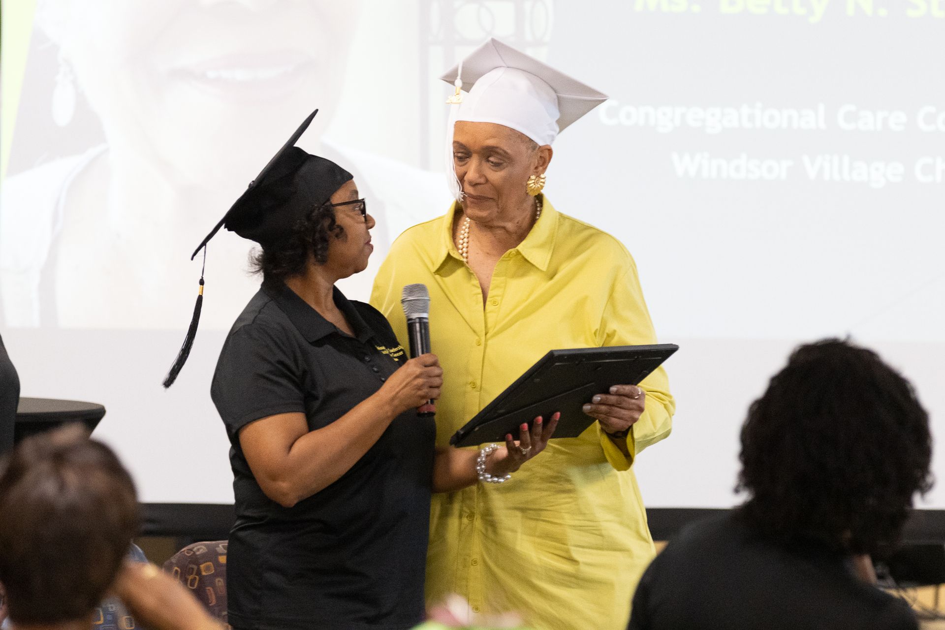 Woman in black shirt speaks into a microphone as another woman in yellow and a graduation cap holds a tablet.