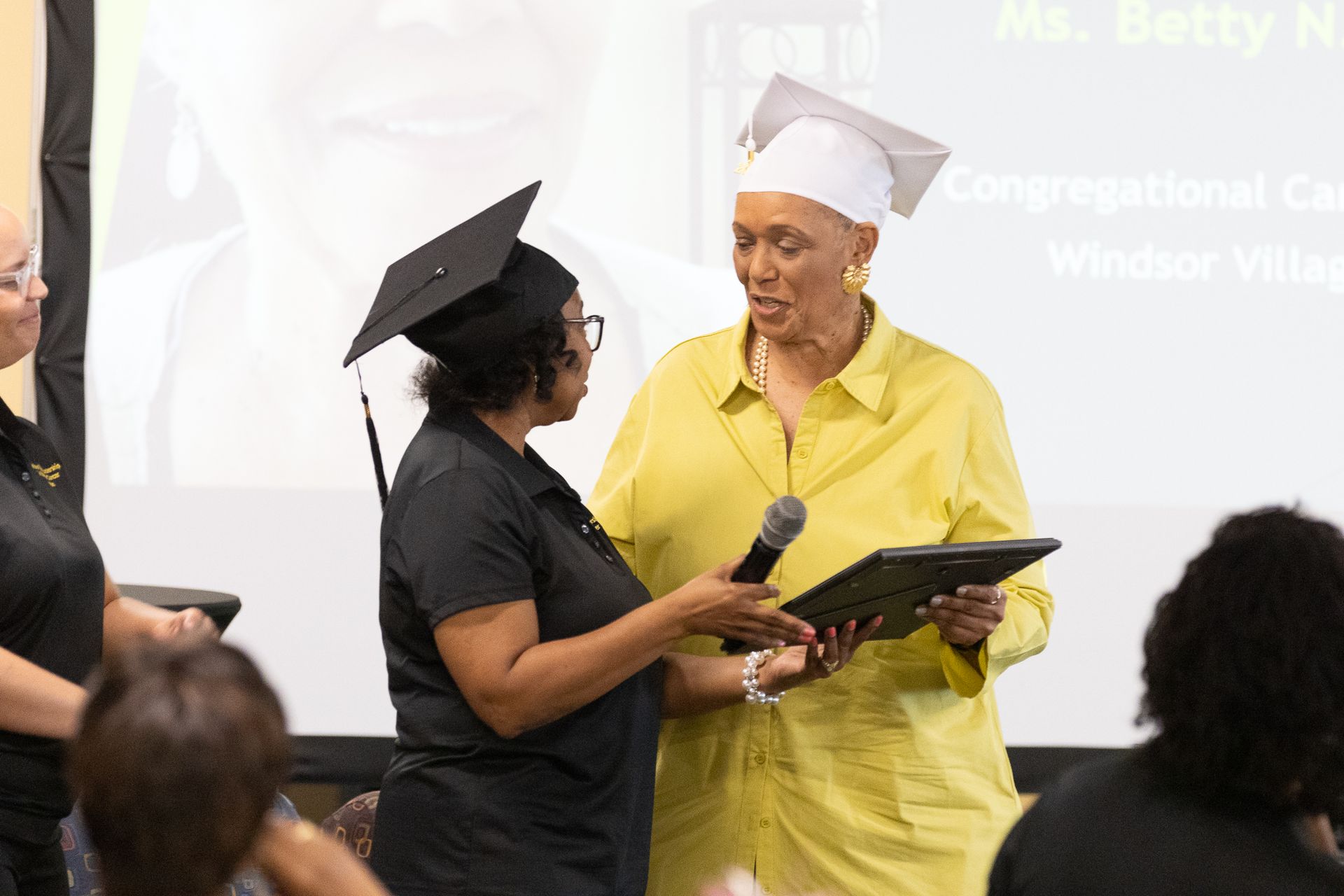 Woman in yellow jacket presents an award to another woman in a graduation cap. Event in a room.