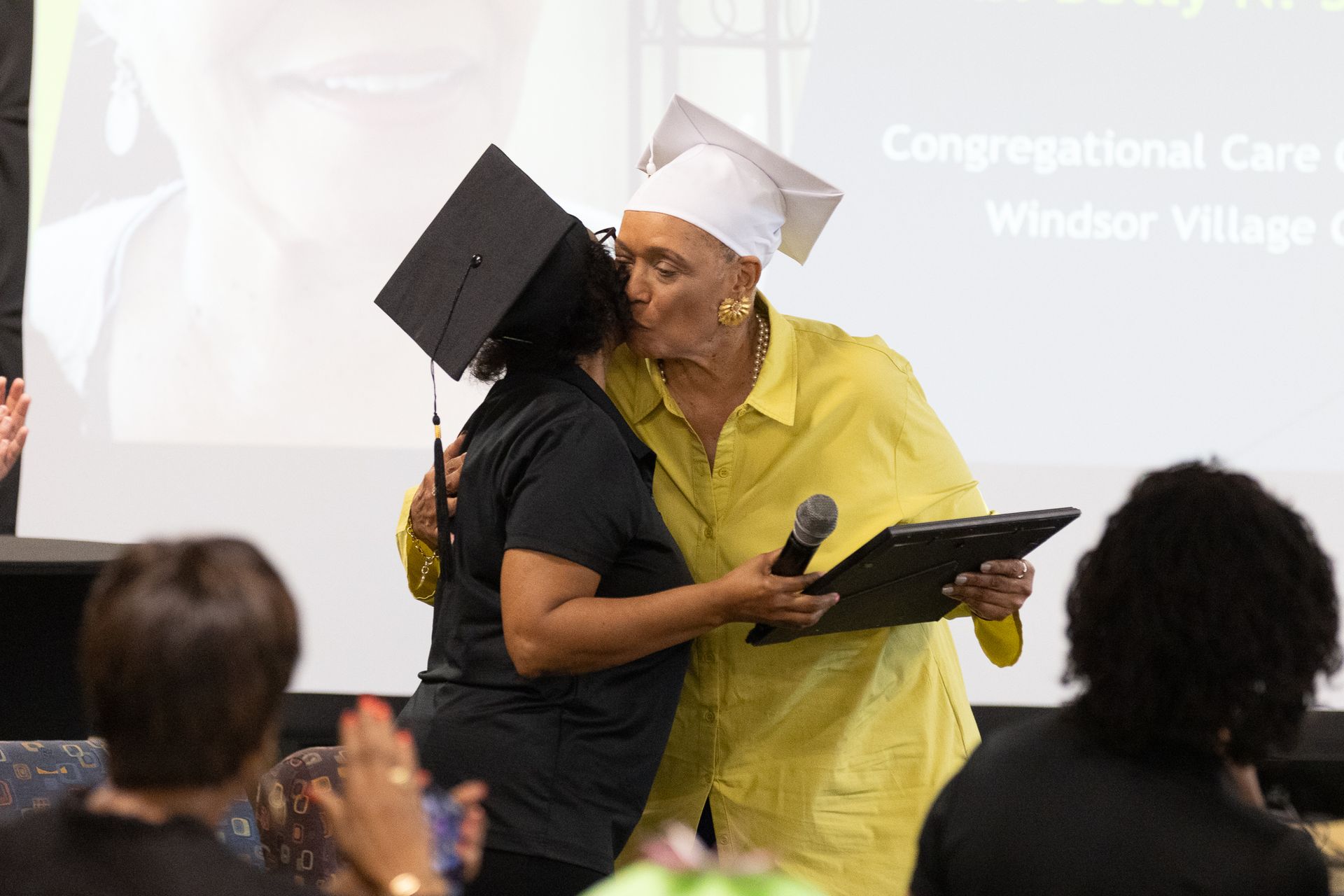 Woman in mortarboard kissed by another in a yellow shirt at an event.