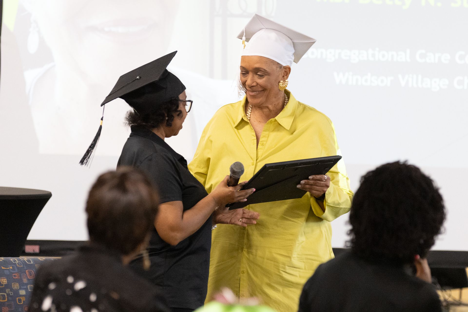 Woman in yellow jacket receives framed document from another woman at an event. Both wear graduation caps.