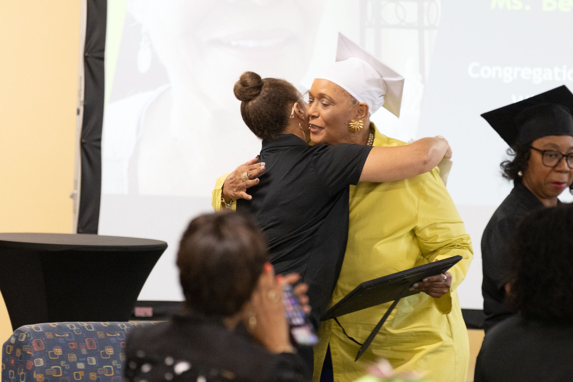 Woman in graduation cap hugs another woman, both smiling, at a ceremony.