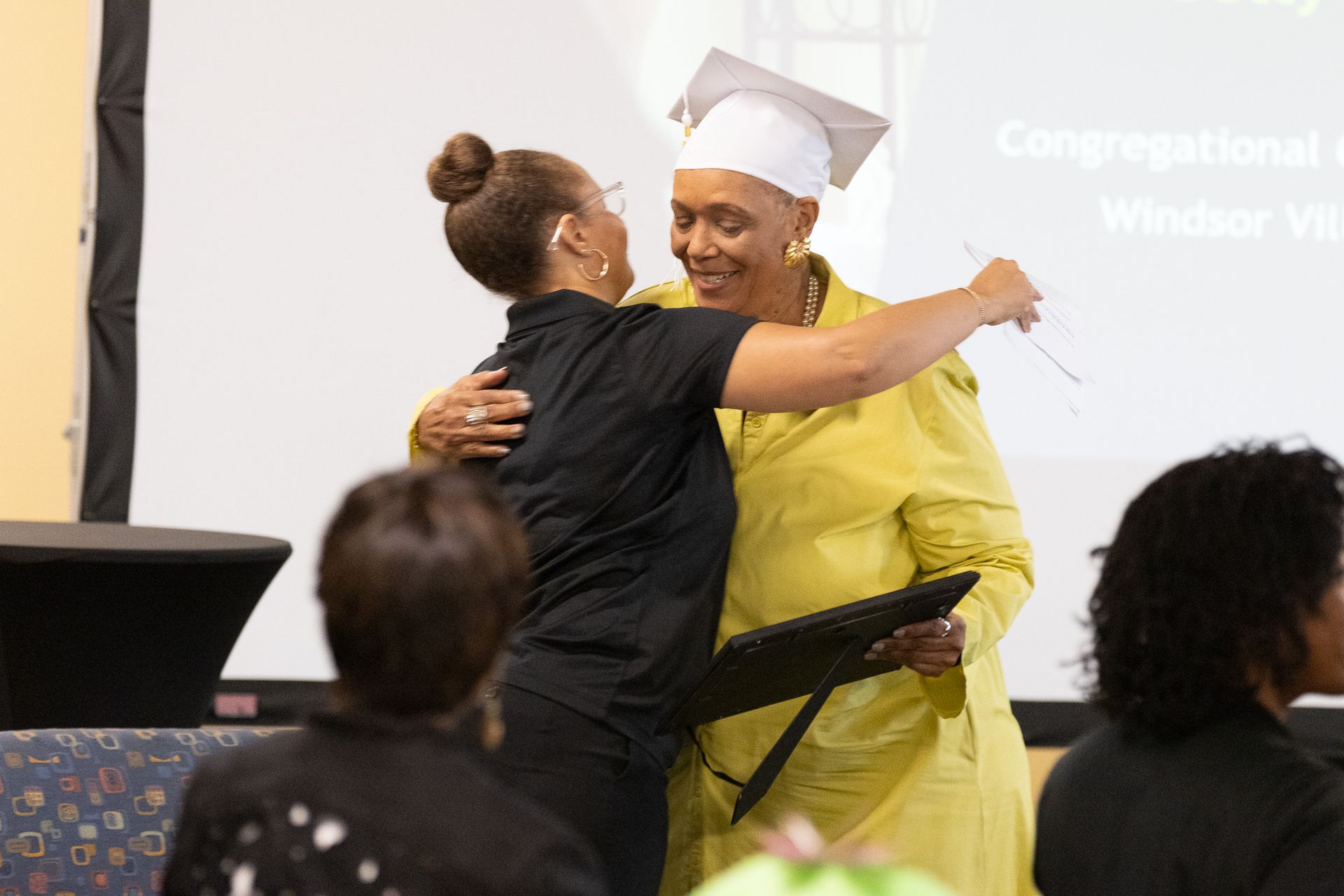Two women embrace at an event. One wears a graduation cap and yellow jacket, holding a document.