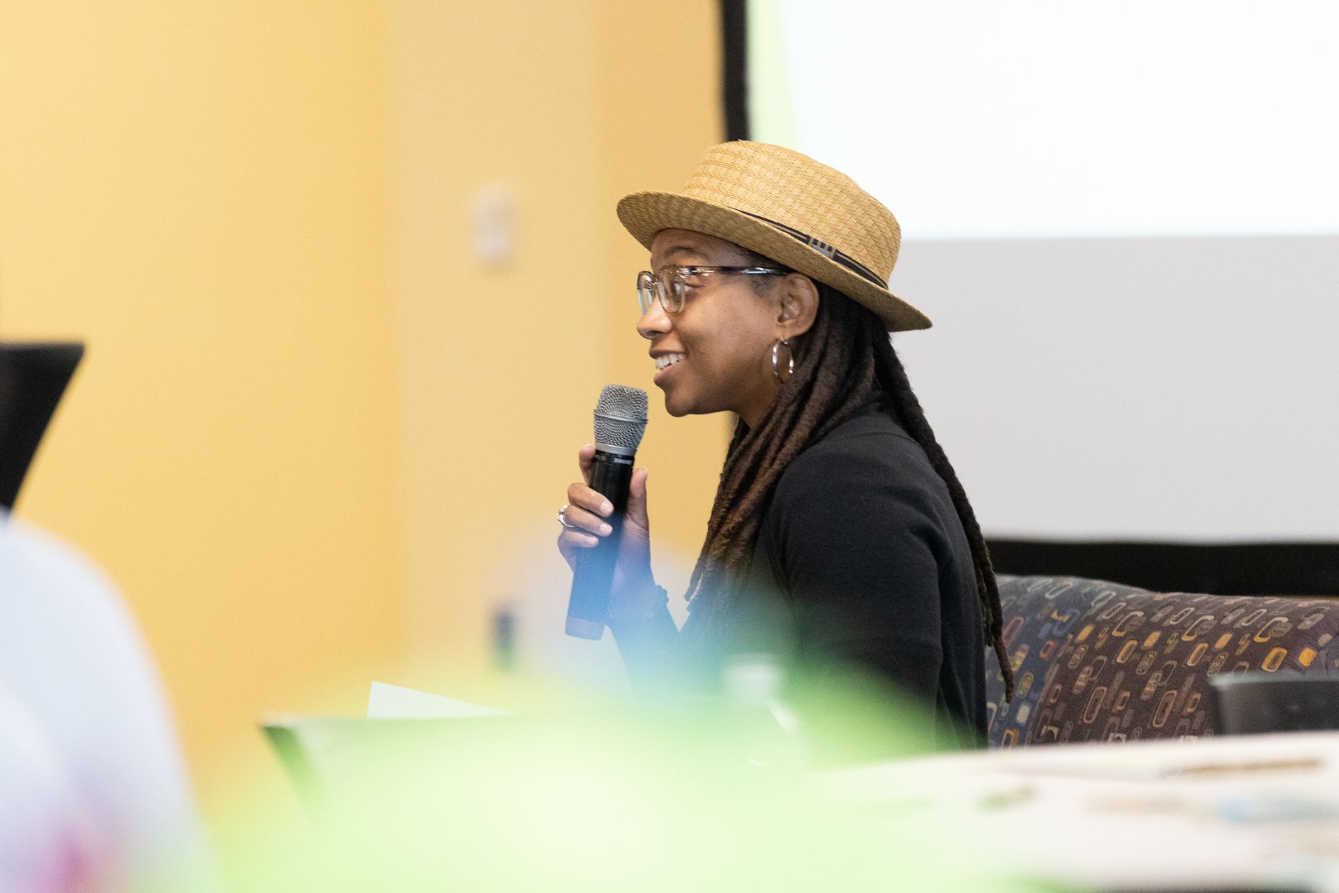 Woman speaking into a microphone, wearing a straw hat and glasses, in front of a white screen, during a presentation.