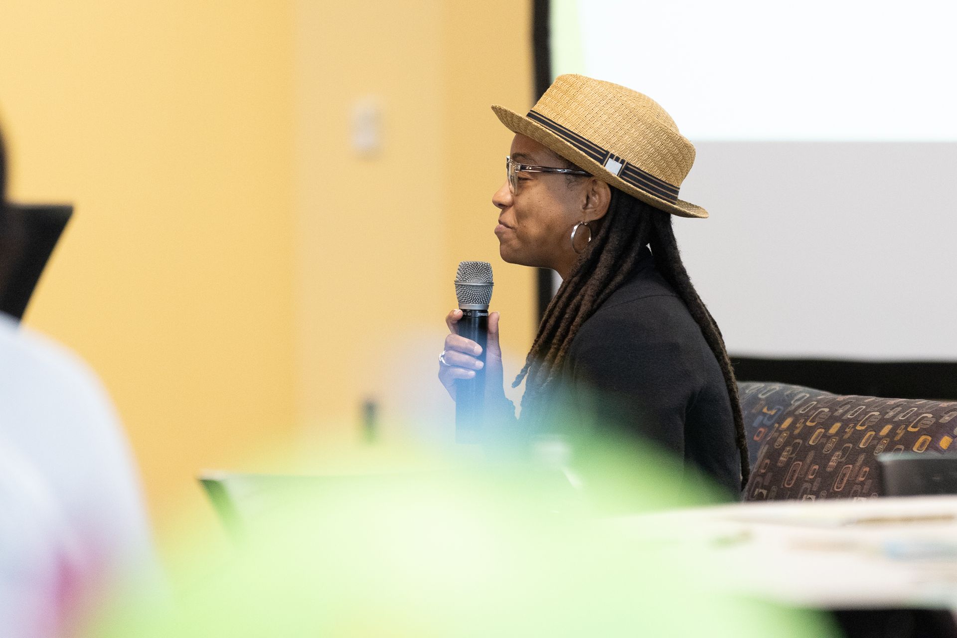 Woman speaking into a microphone, wearing a straw hat and glasses, in a conference setting.