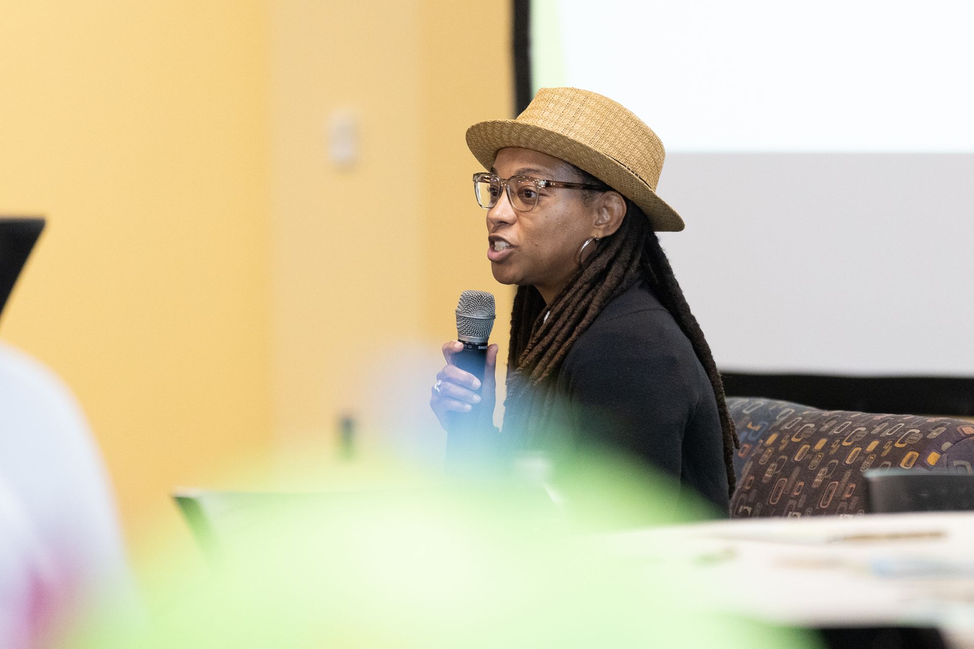 Woman in a straw hat speaks into a microphone at a conference, blurred foreground.