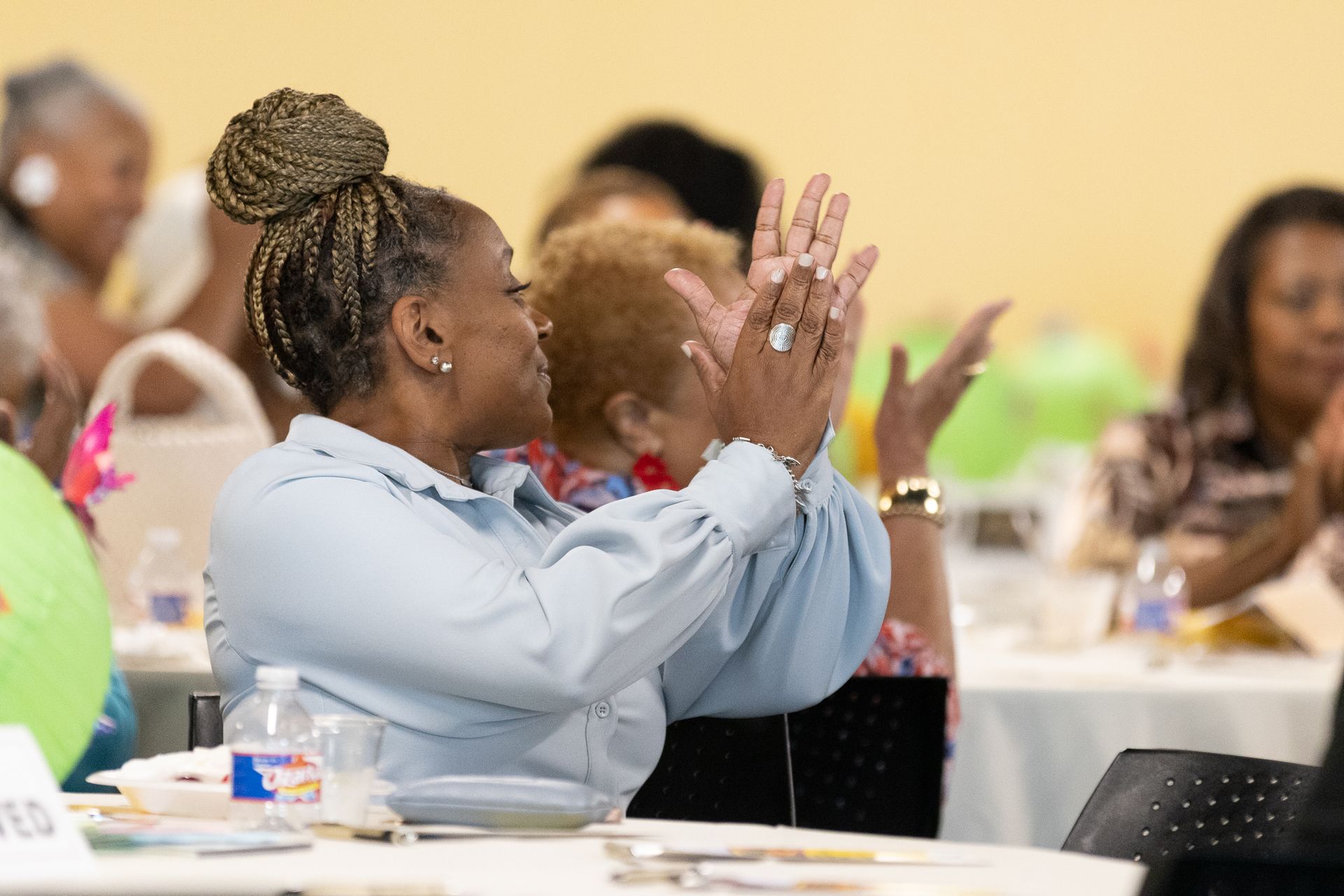 Woman in blue shirt clapping at an event. Other people at tables in the background.