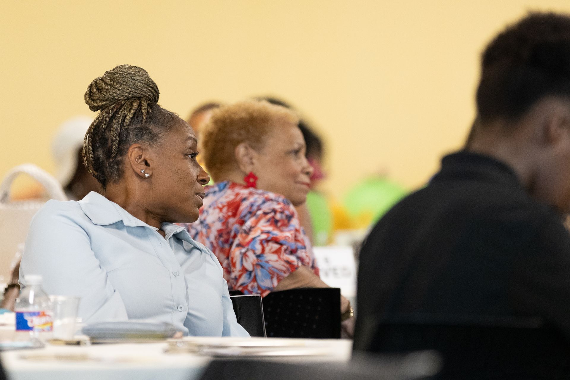 People seated at tables in a conference room, listening attentively. Woman with updo looks to the side.