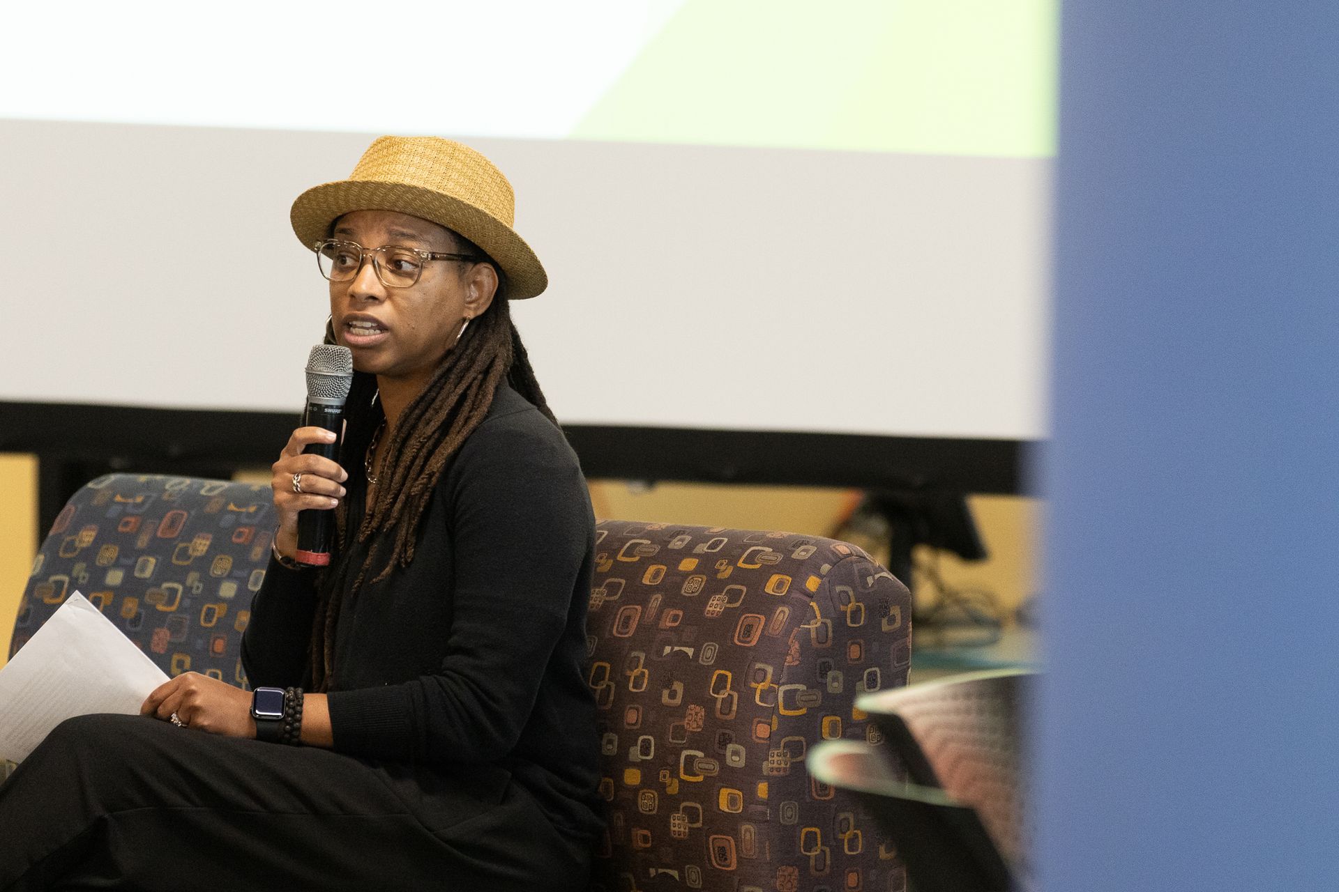 Person with dreadlocks, in a straw hat, speaking into a microphone while seated; holding papers.