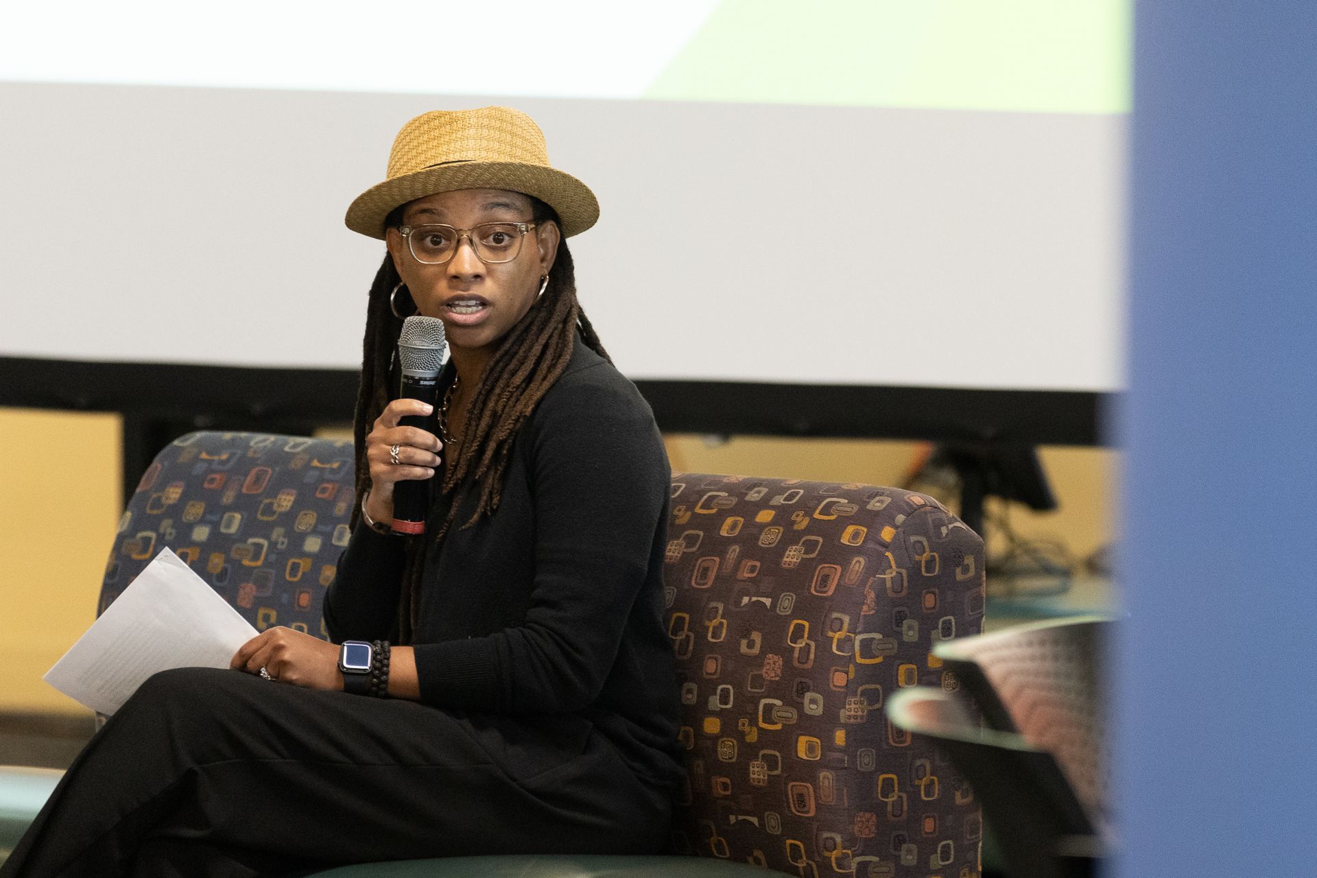 Woman in hat and glasses speaks into a microphone, holding papers, seated at an event.