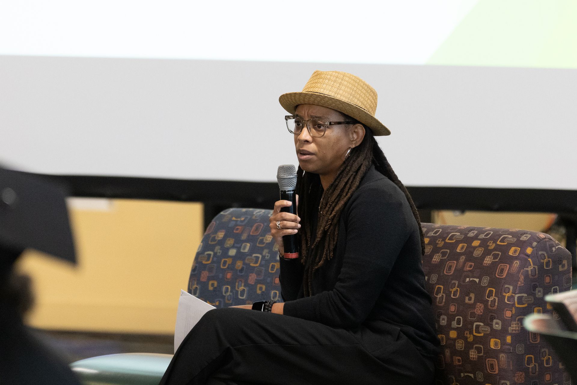 Person wearing a hat and glasses speaking into a microphone while seated on a patterned chair.