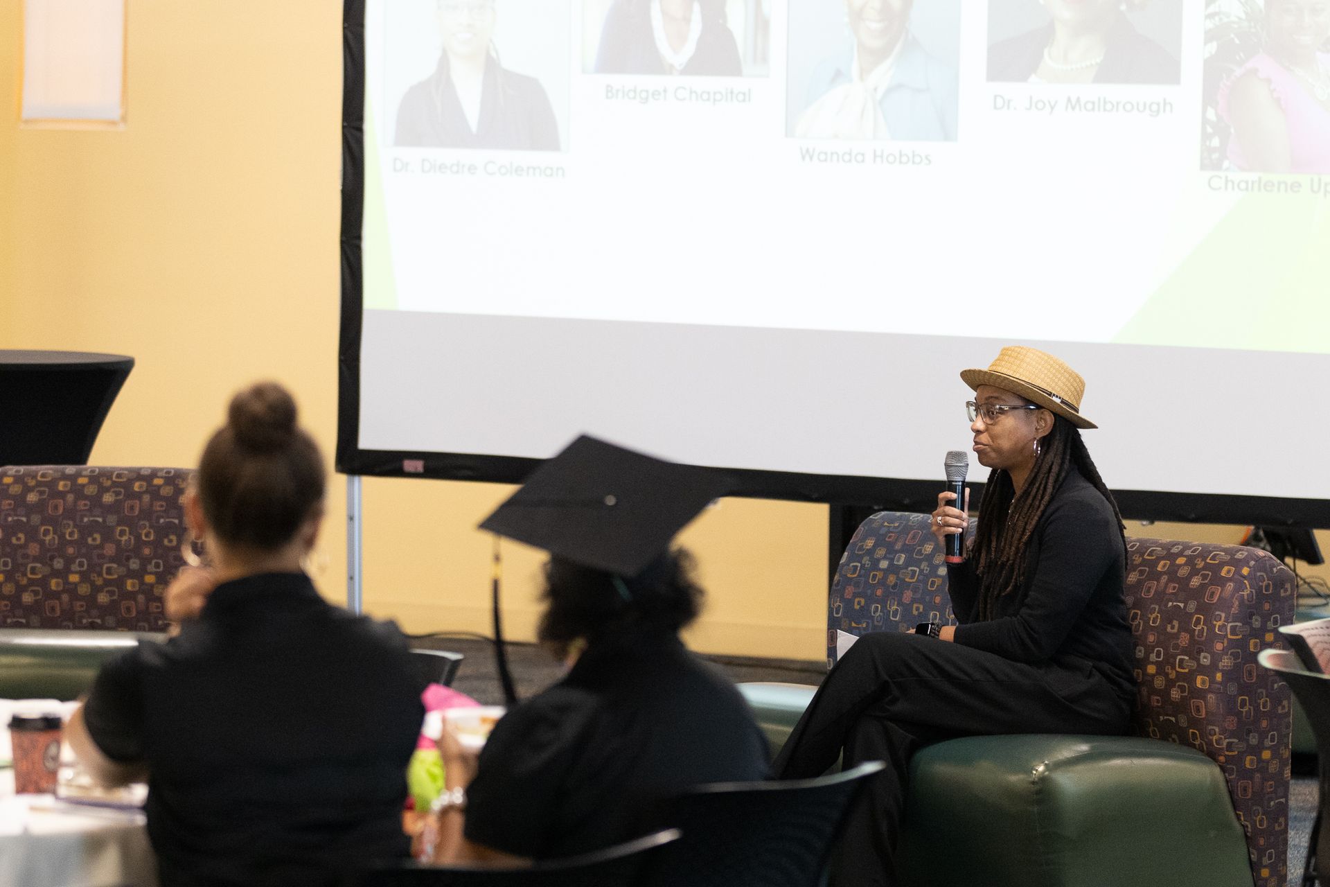 Woman speaking into a microphone during an event, in front of a screen displaying names. Attendees in the foreground.