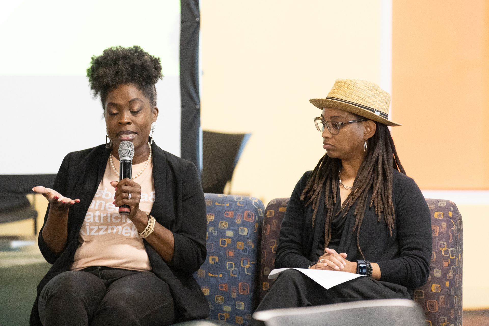 Two people seated, one speaking into a microphone. They are participating in a panel discussion.