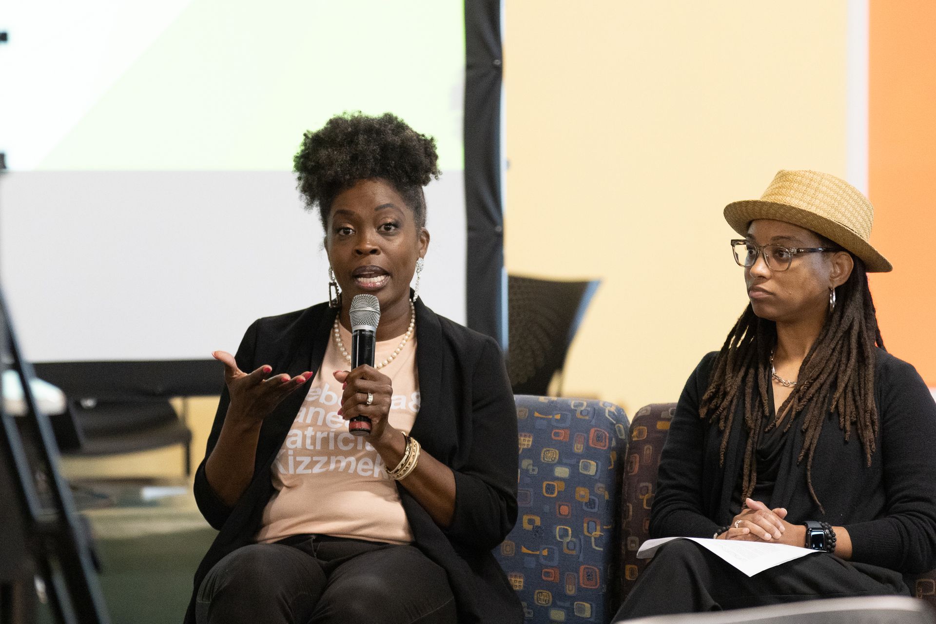 Two women on a panel, one speaking into a microphone, gesturing, other listening, wearing a hat.