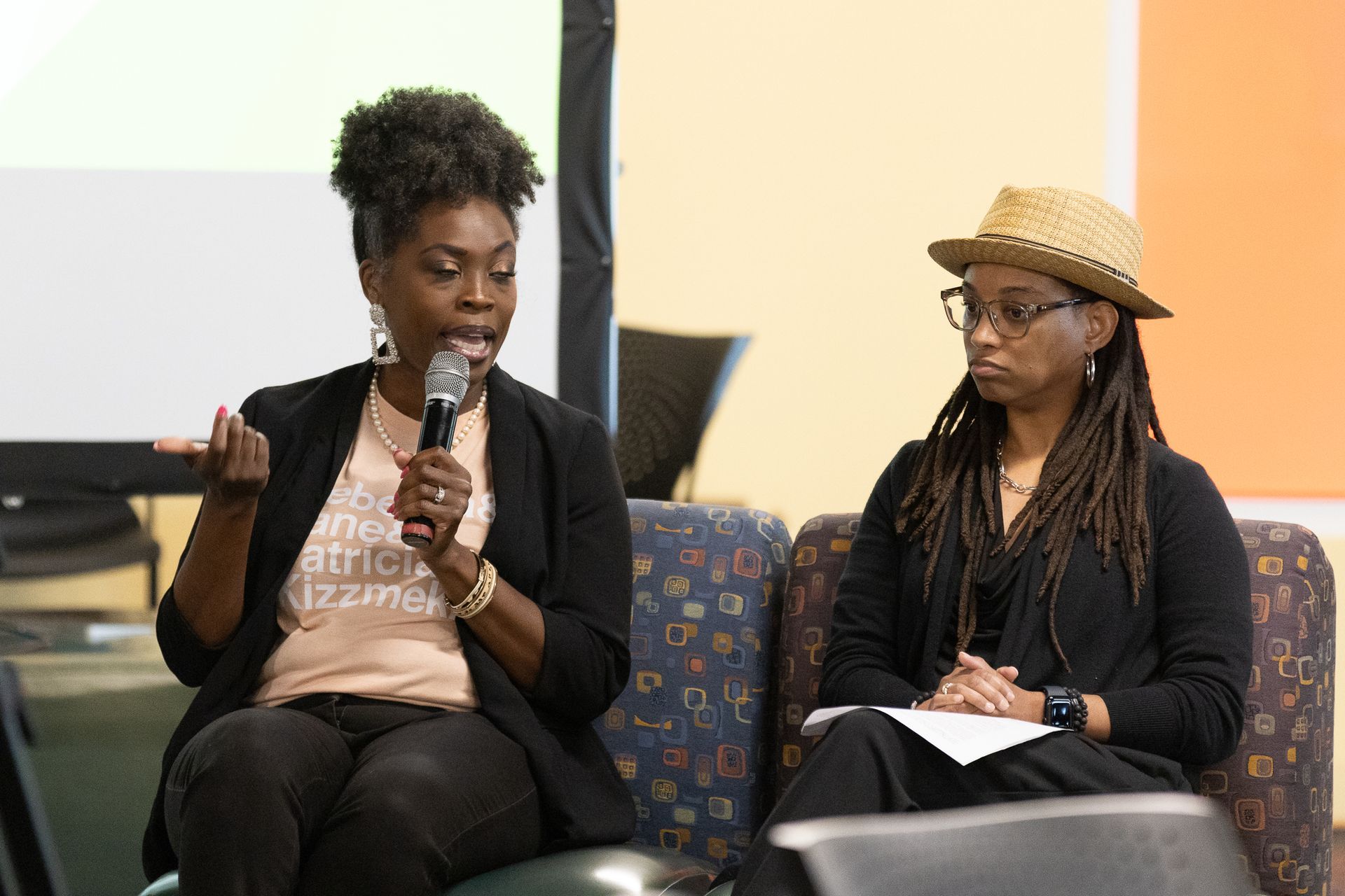 Two women seated, one speaking into a microphone. Panel setting. Orange and green background.