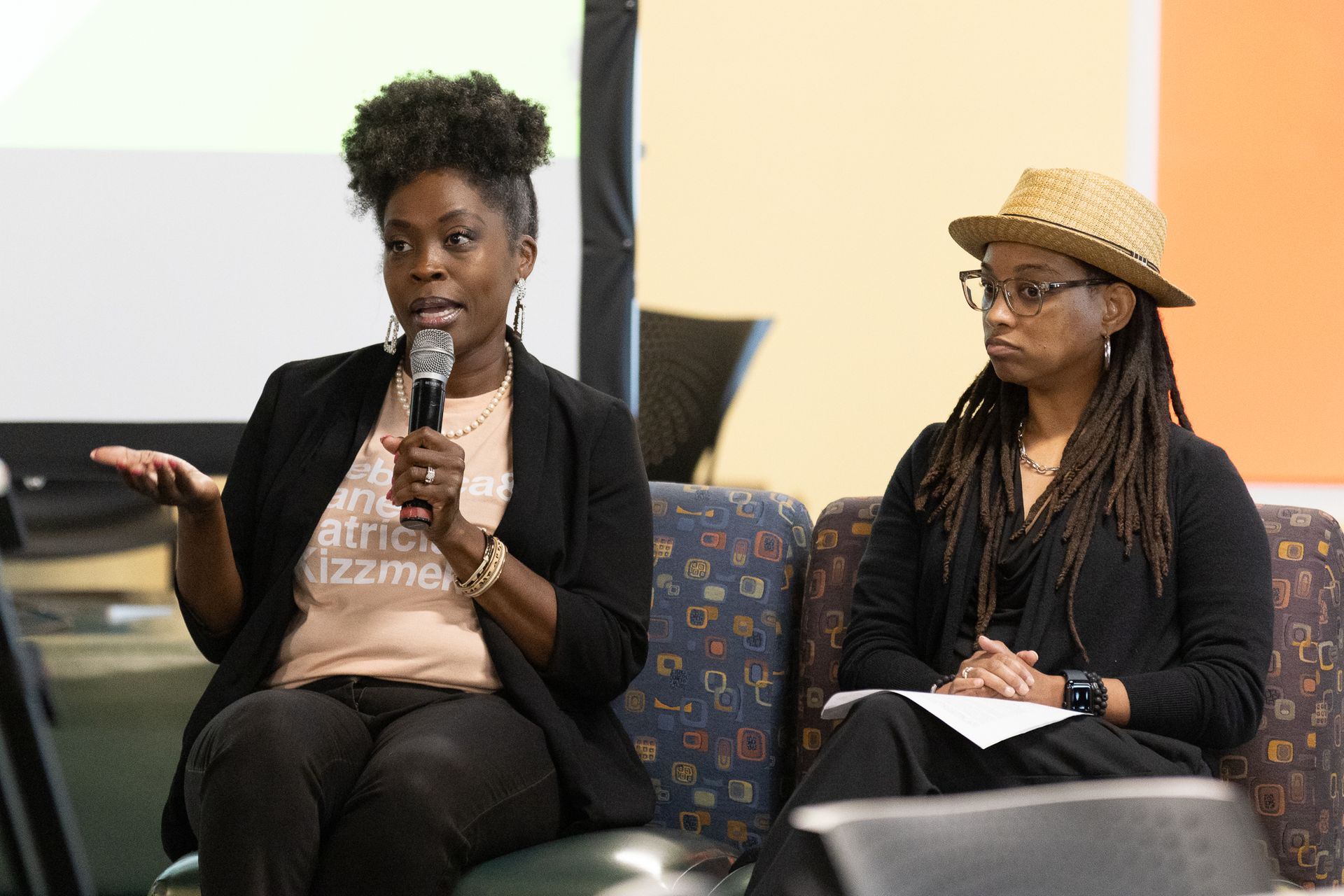 Two people seated, one speaking into a microphone. They are on stage, with a projector screen behind them.