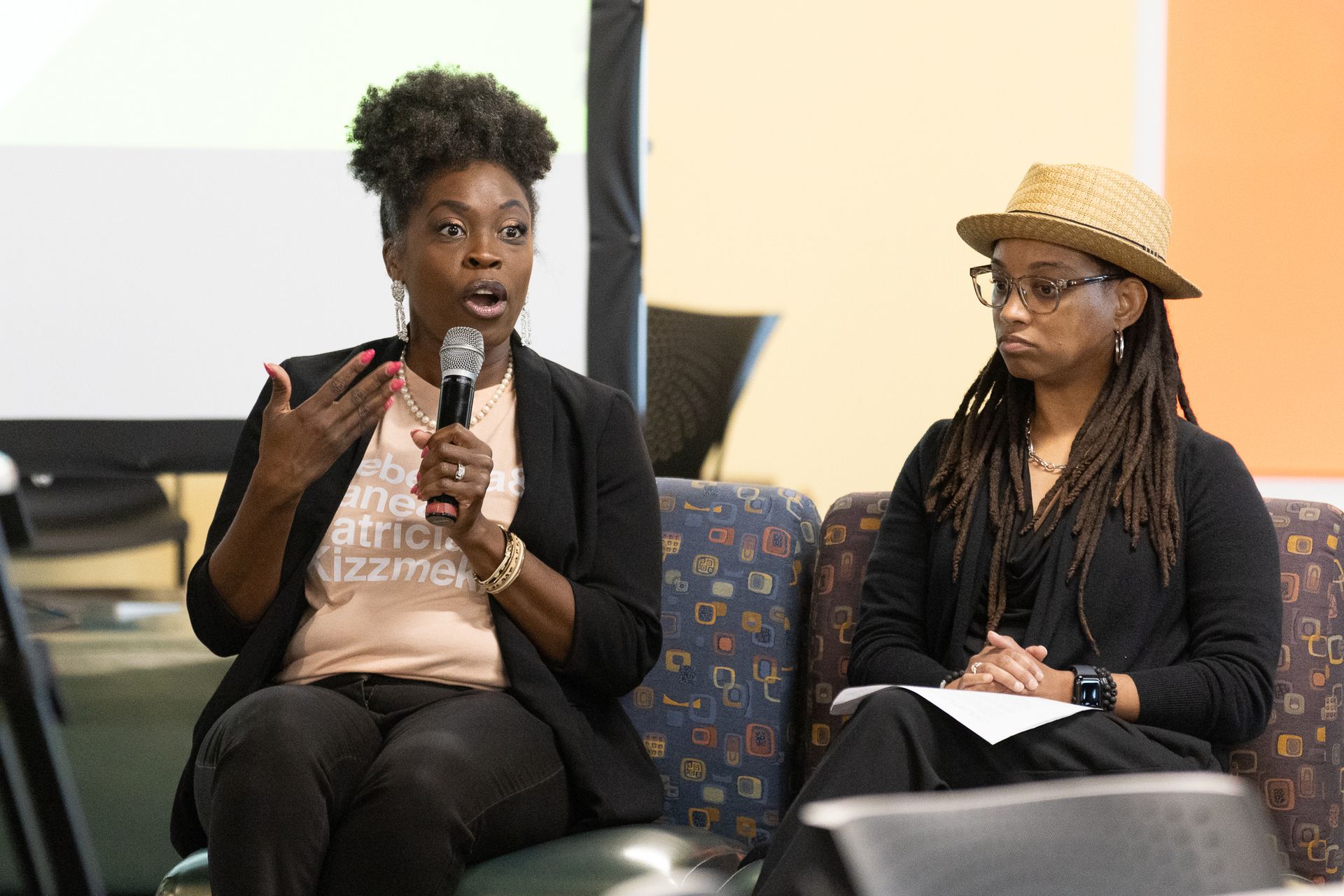 Two people seated at a panel discussion. One speaks into a microphone, gesturing; the other looks on.