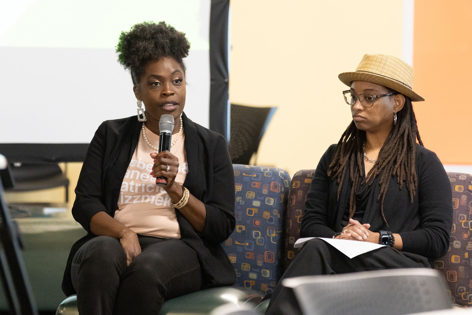 Two people at a panel: one speaks into a mic, the other looks on, holding a paper.