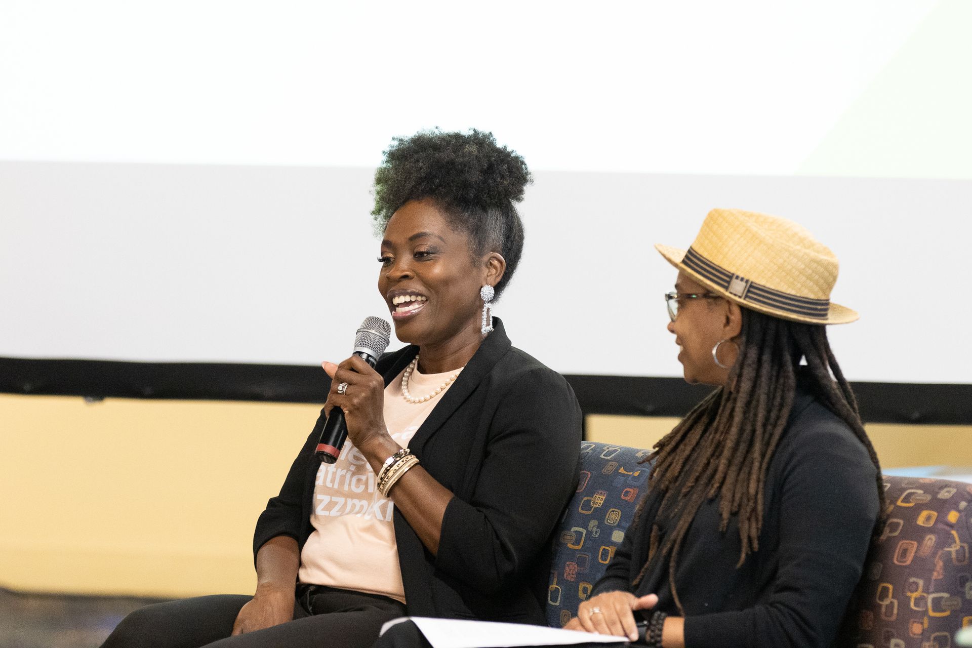 Two women seated at an event, one speaking into a microphone, the other listening.
