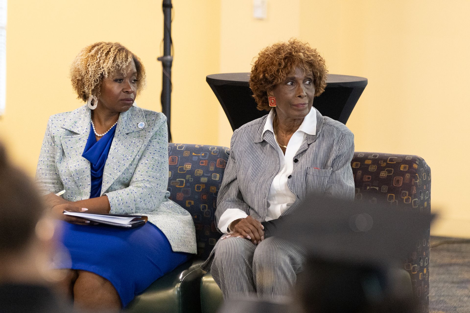 Two women in business attire seated, listening attentively. One holds a pad, the other looks off-camera.