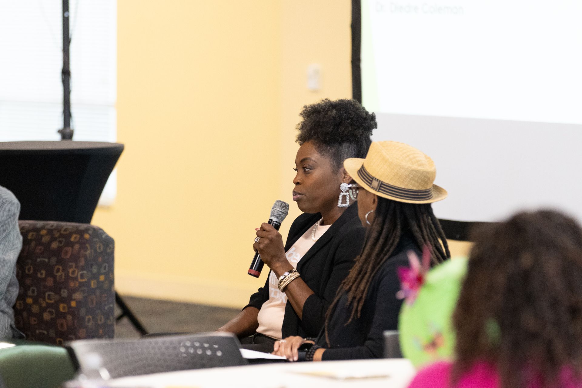 Two women at a table, one speaking into a microphone. Both are wearing blazers; one has a hat. Bright room.