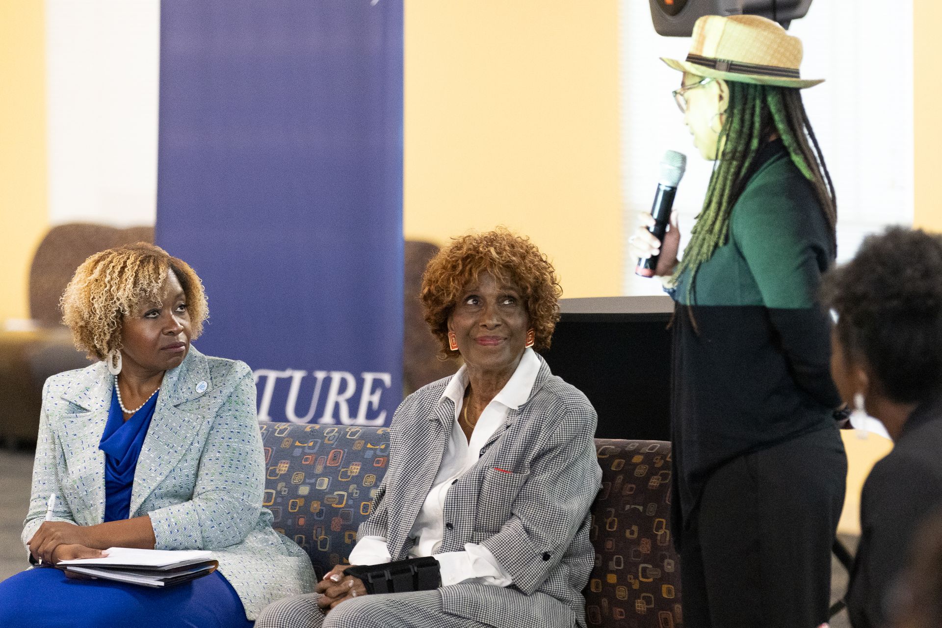 Two women seated listen to a speaker holding a microphone; event setting.