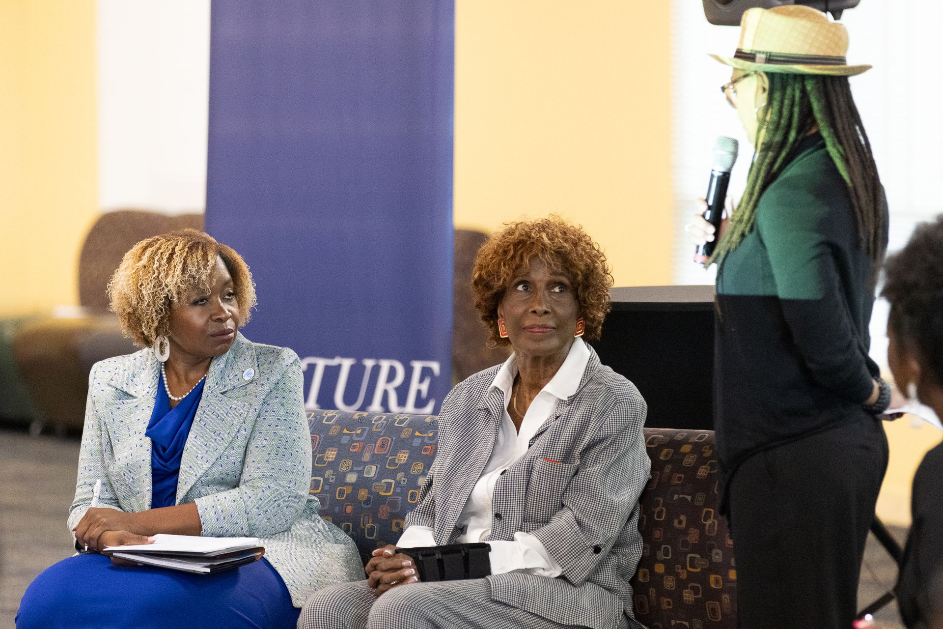Three people seated during an event; one speaks into a microphone.
