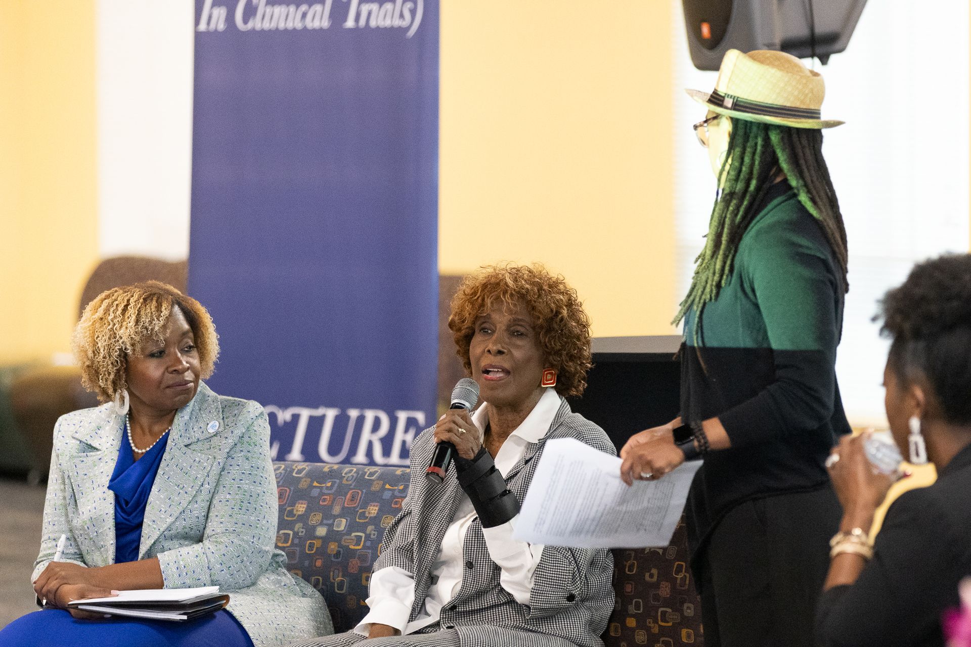 Three women on stage during a lecture. One holds a microphone, speaking. The setting is indoors.