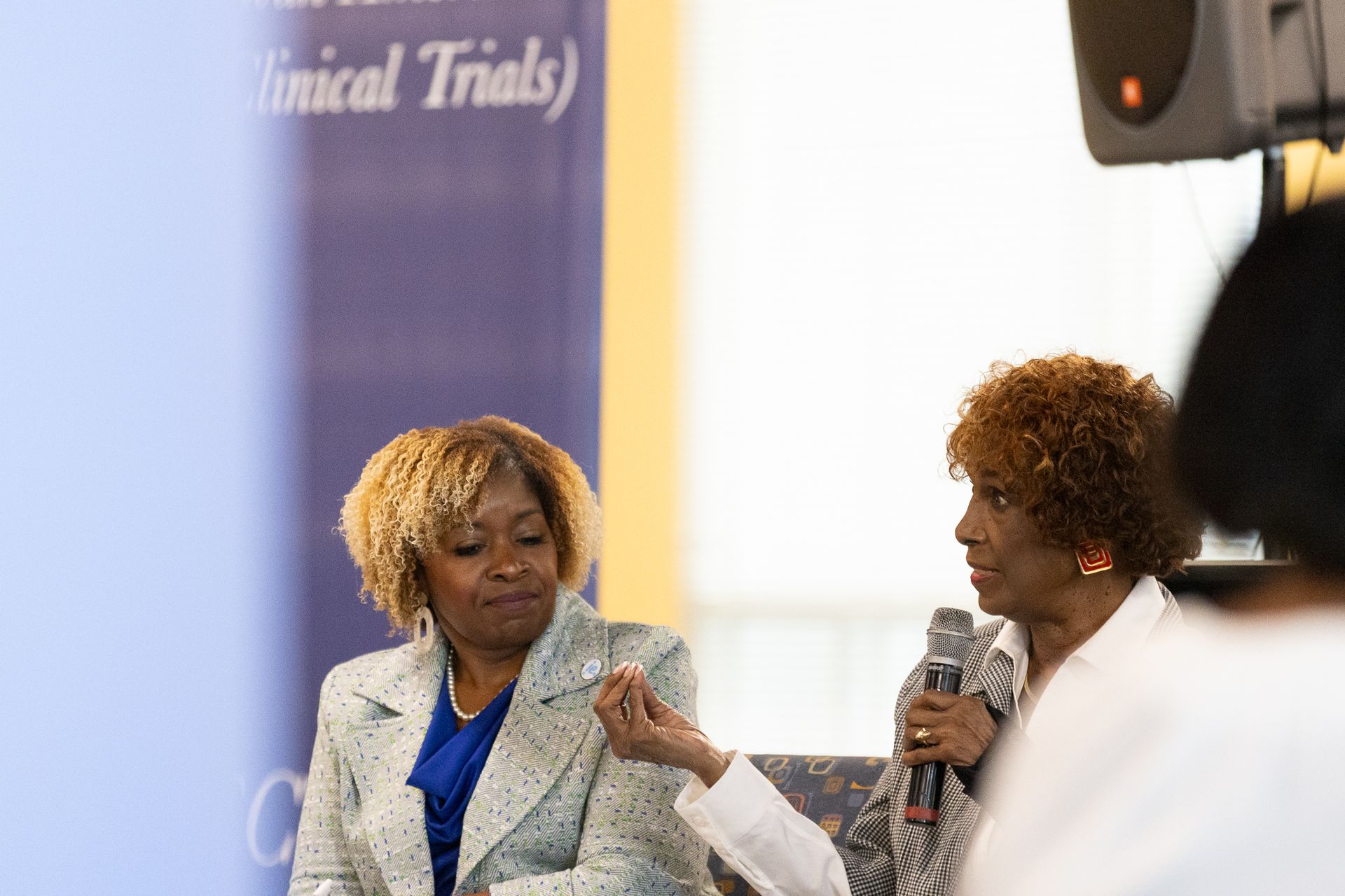 Two women at a panel discussion. One speaks into a microphone, gesturing. The other looks on.