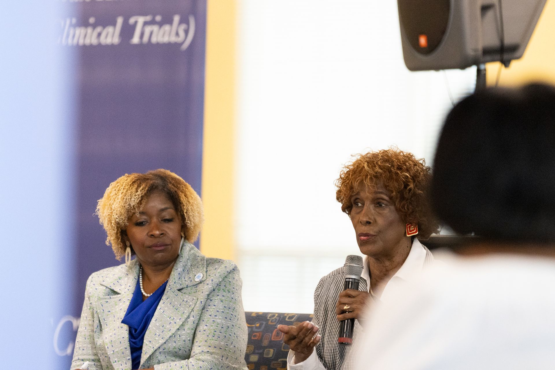 Two women at a panel discussion; one speaks into a microphone. Banner in background.