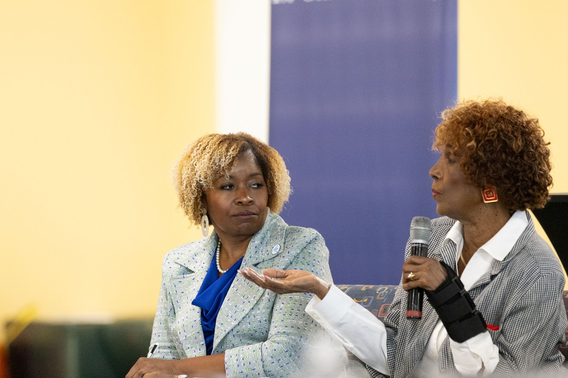 Two women at a panel discussion; one speaks into a microphone, gesturing, the other looks on.
