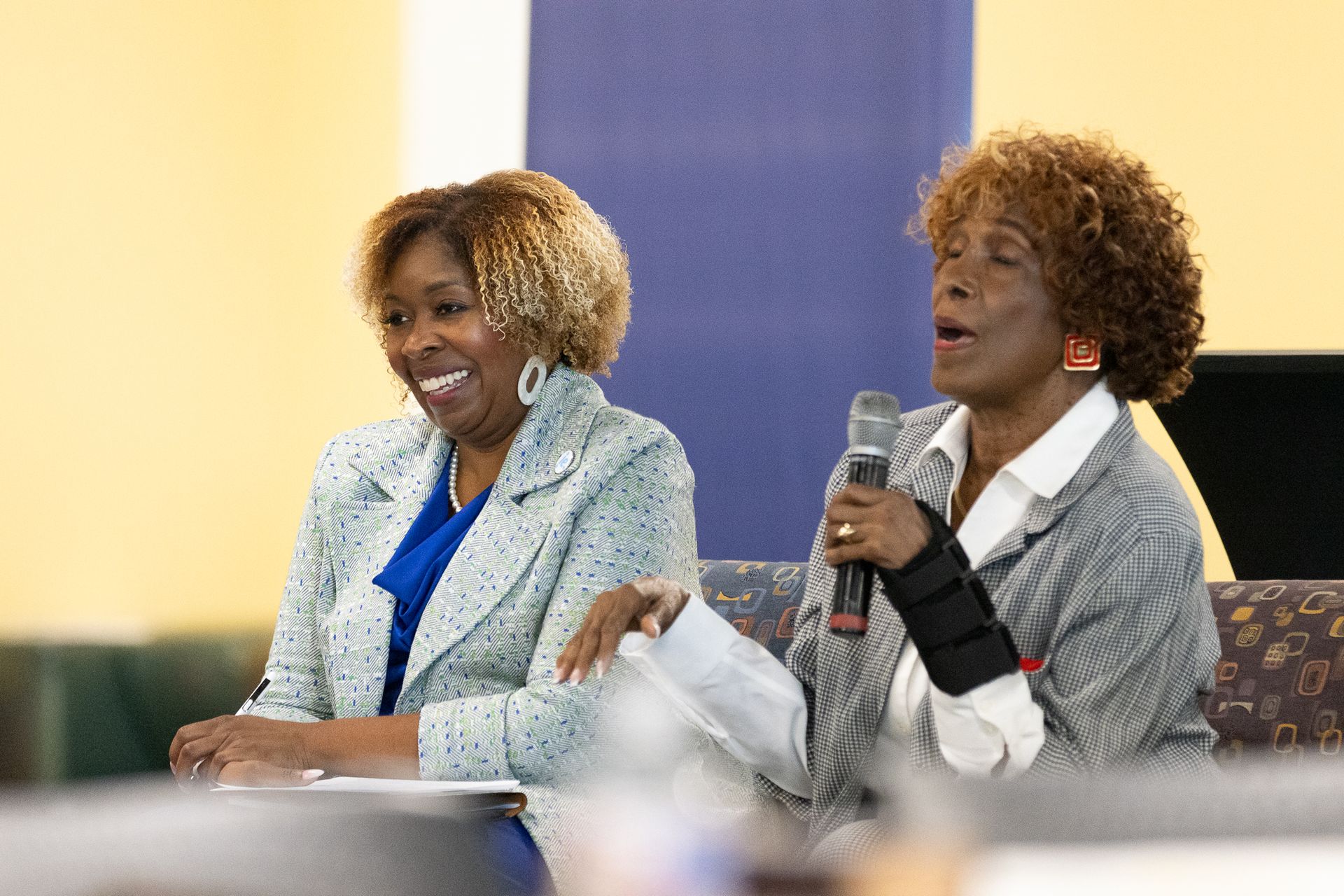 Two women at a table, one speaking into a microphone. Both are smiling, one wearing a brace on her arm.