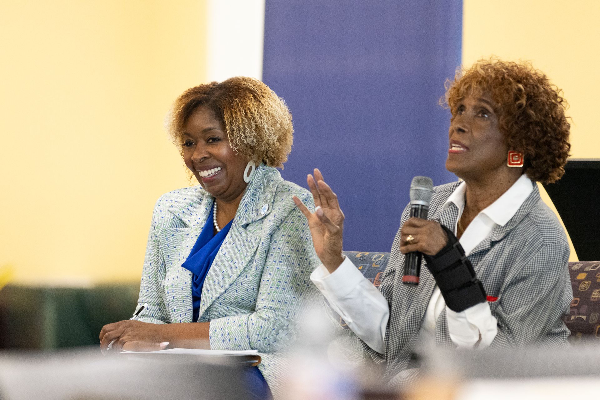 Two women seated, one speaking into a microphone. Both are smiling, wearing jackets, and seated indoors.