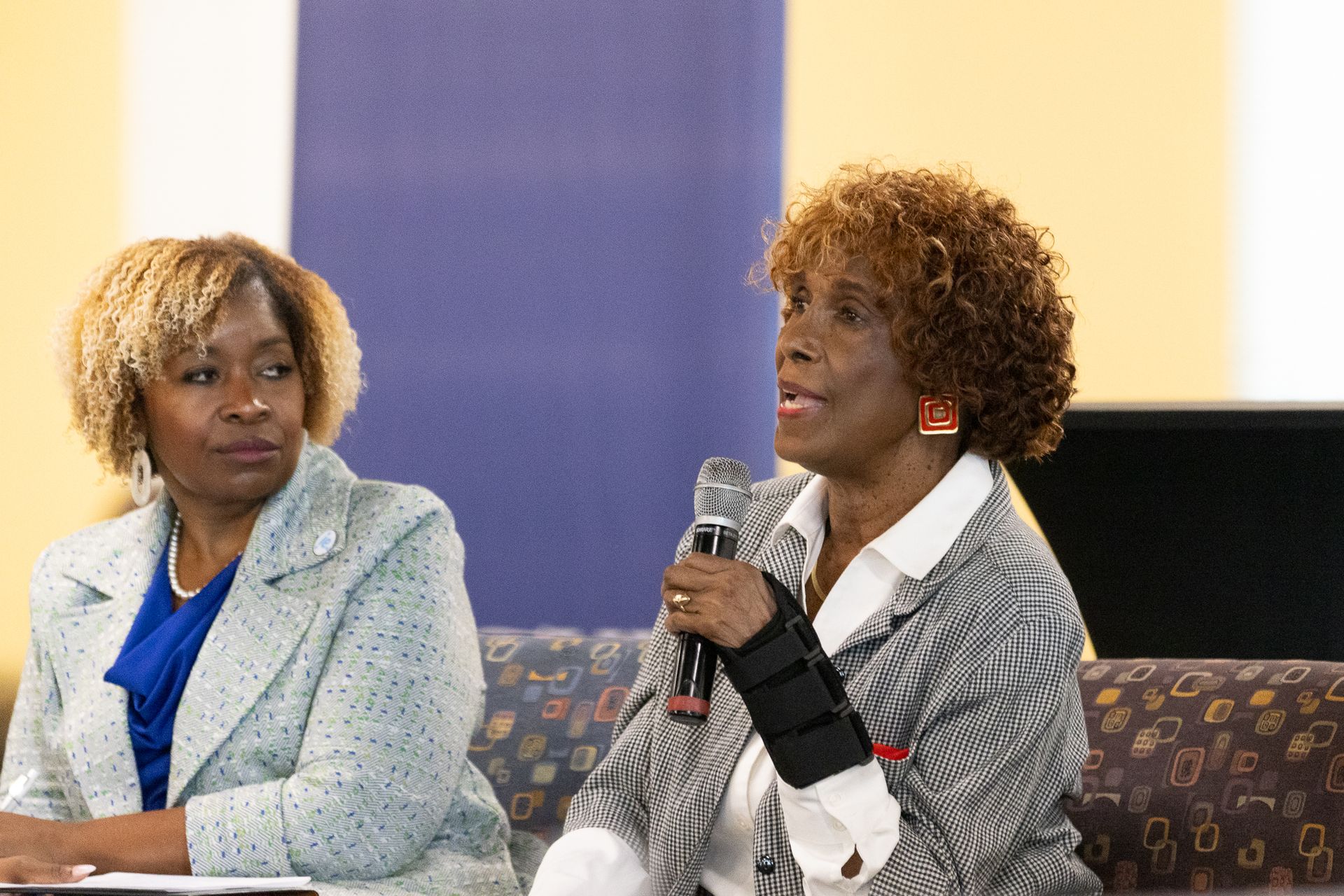 Two women at a table, one speaking into a microphone with arm brace; backdrop is purple and yellow.