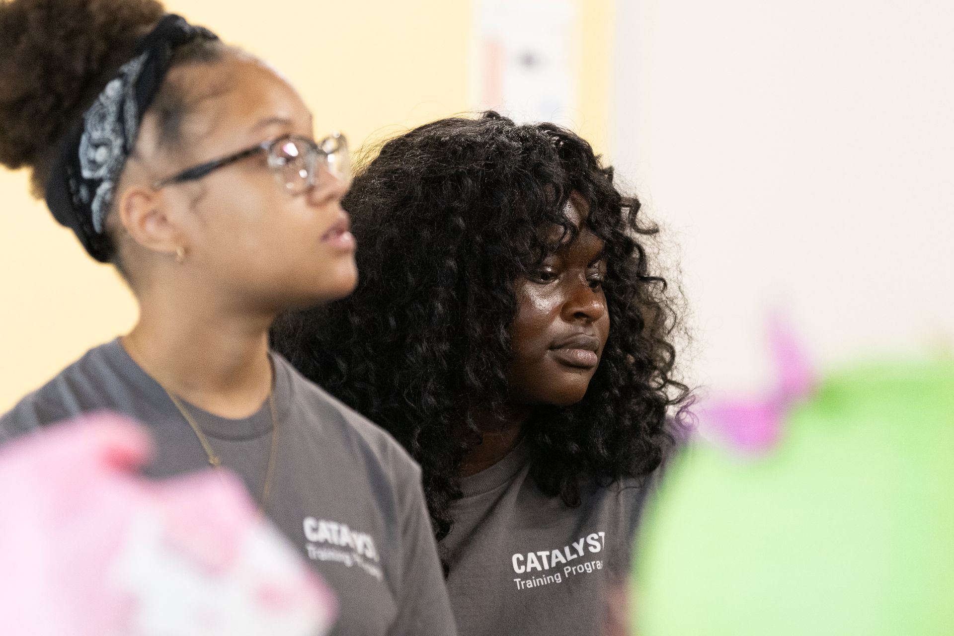 Two young people in gray shirts, looking attentively to the right. One has a headscarf, glasses; the other has curly hair.