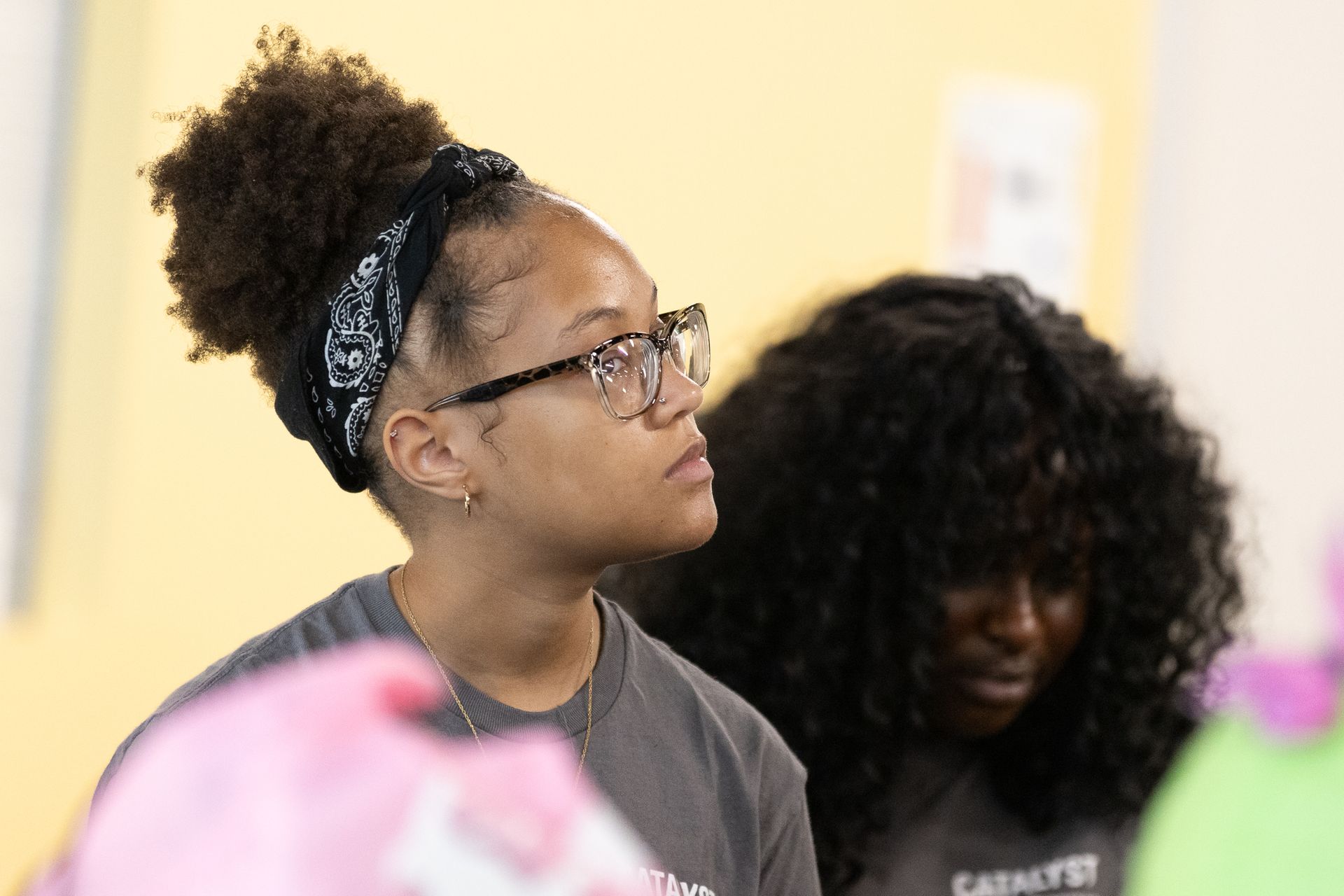 Woman wearing glasses and a bandana looks upward; another woman with curly hair is partially visible.