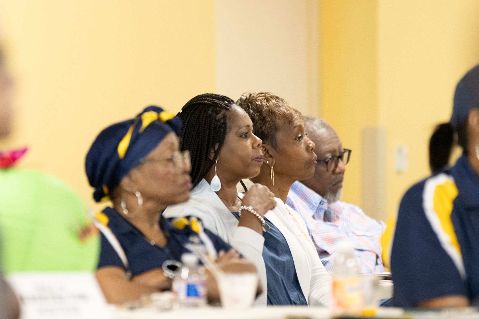 People seated at tables, listening attentively. Yellow wall in background.