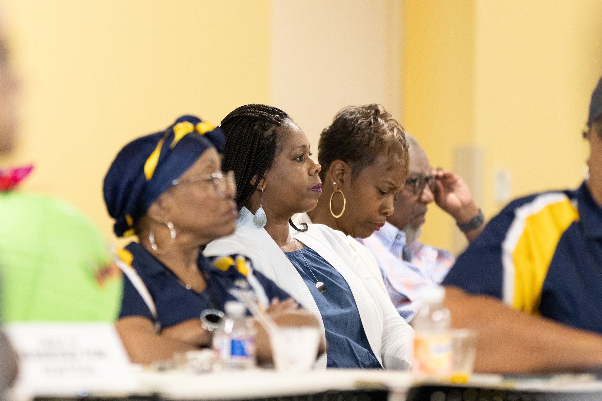 People seated at a table, some looking forward, others looking down. Yellow and blue clothing, glasses.